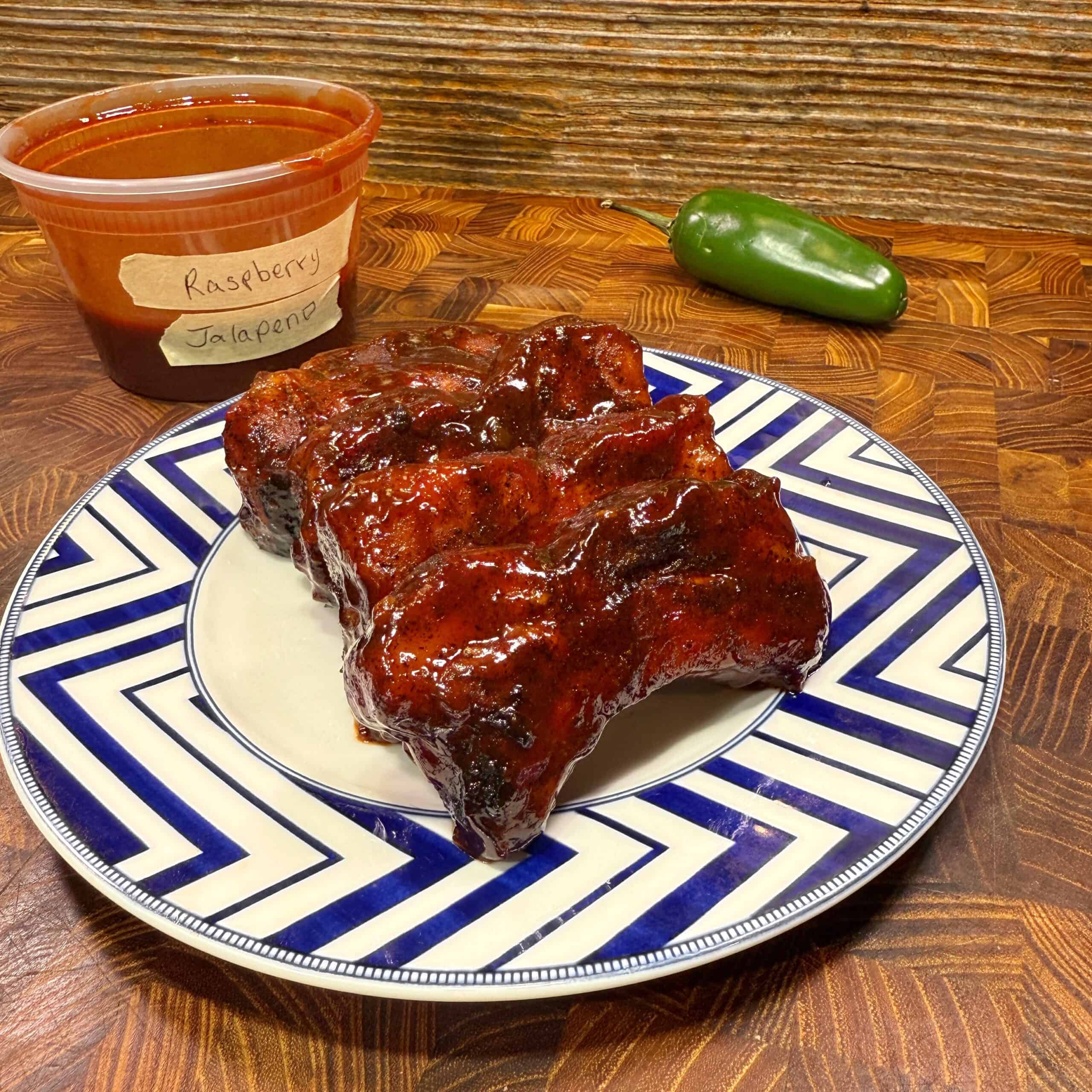 Raspberry Jalapeno Party Ribs ribs coated in a glossy sauce are served on a blue and white patterned plate. Behind them is a container labeled “Raspberry Jalapeno” and a whole jalapeño pepper on a wooden surface.