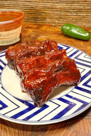 A plate of glazed ribs sits on a zigzag-patterned plate, with a container labeled Raspberry Jalapeno, hinting at the delicious Raspberry Jalapeno Party Ribs Recipe, and a fresh jalapeno pepper in the background on a wooden surface.