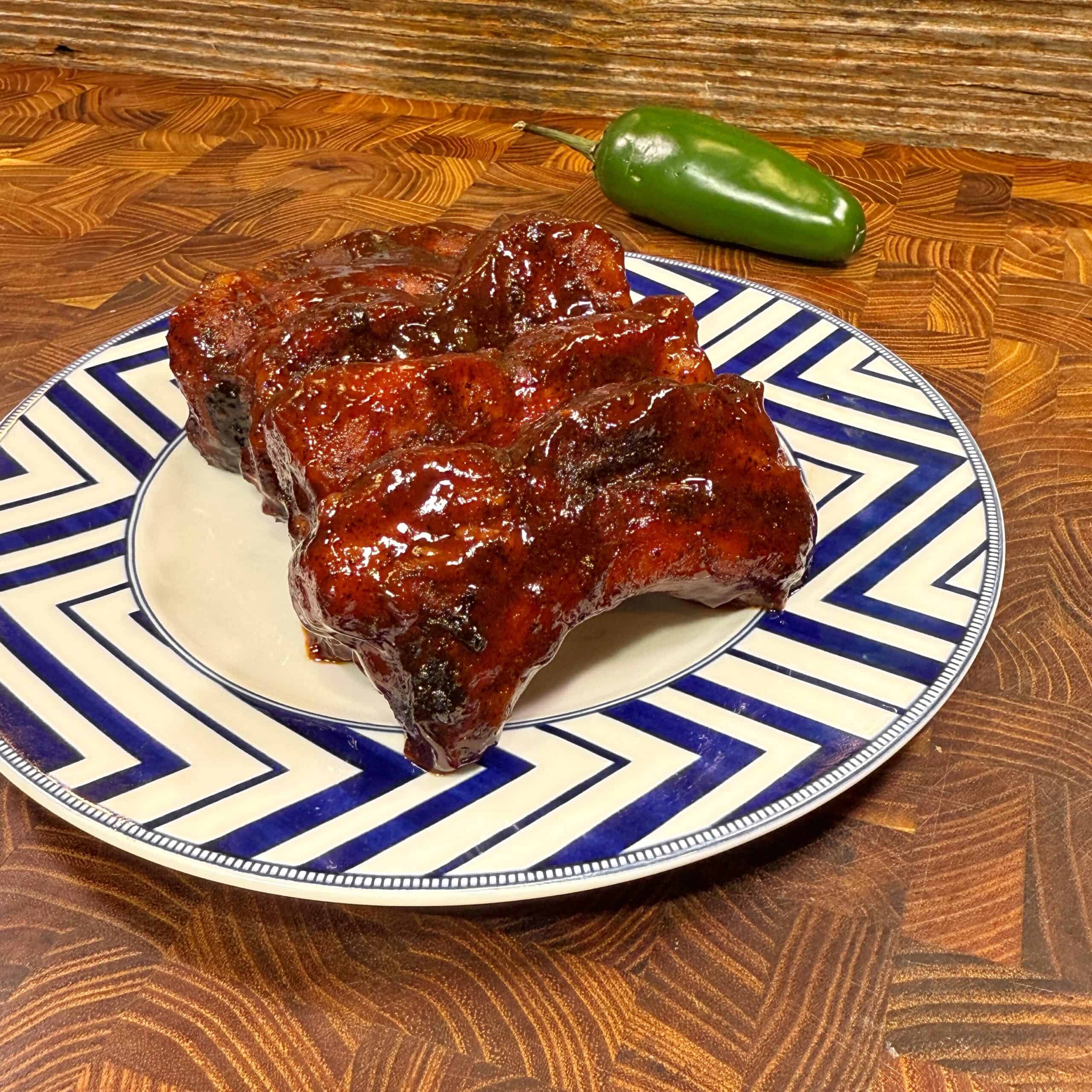 Barbecued ribs coated in a shiny glaze are stacked on a patterned blue and white plate, with a whole jalapeño pepper in the background on a wooden surface.