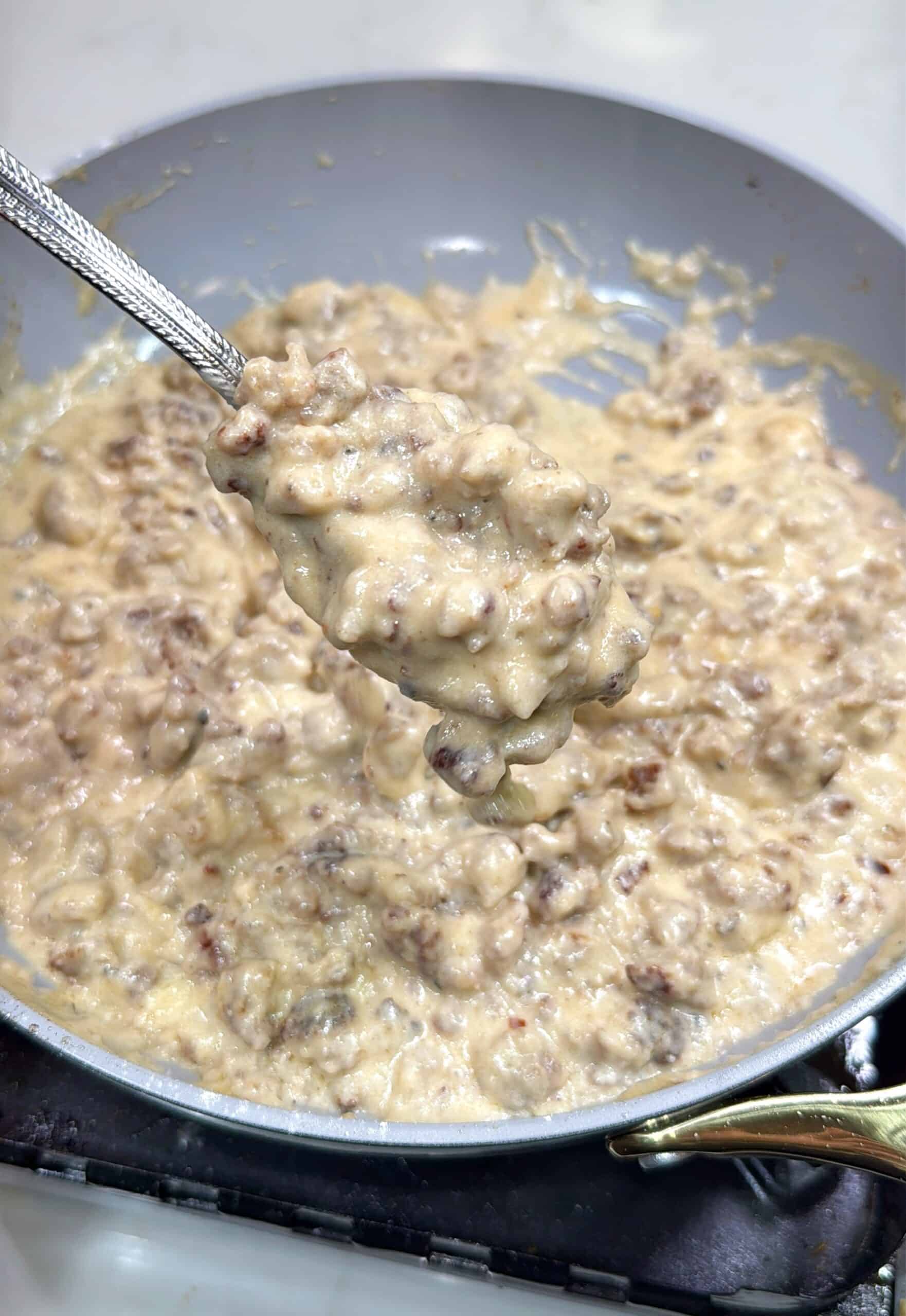 A close-up of a spoonful of creamy sausage gravy being held over a pan filled with more sausage gravy on a stovetop.