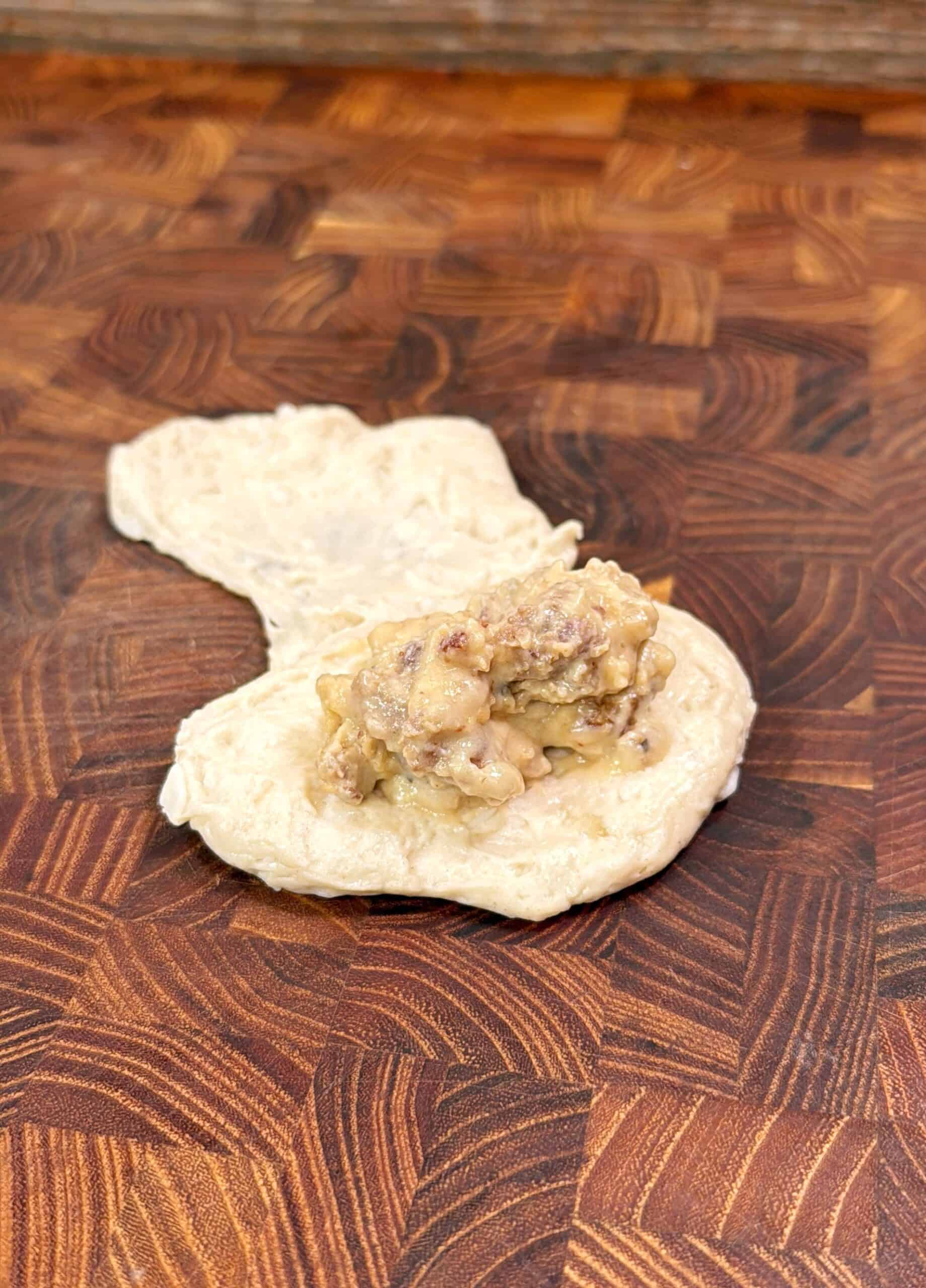 A small portion of dough with a savory meat and gravy filling sits on a wooden cutting board, ready to be folded or shaped for cooking.
