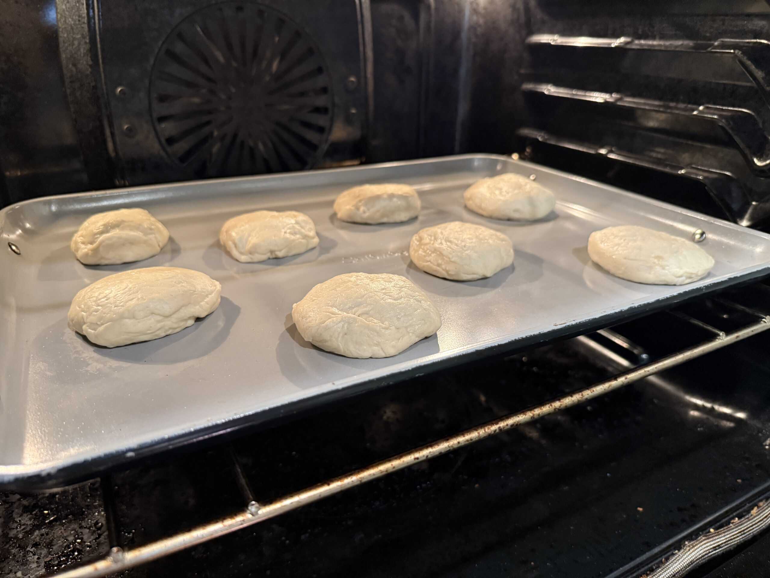 A baking tray with eight raw biscuit dough pieces is placed in an oven, with the oven racks and fan visible in the background.