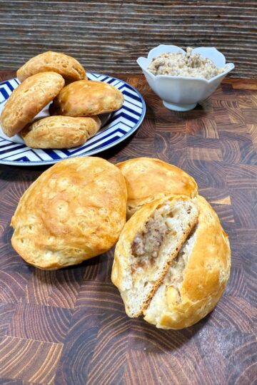 Golden brown bread rolls filled with sausage and cheese are displayed on a wooden surface. Some are on a blue and white plate, a few are in front, with one split open to show the filling. A bowl of sausage mixture sits in the background.