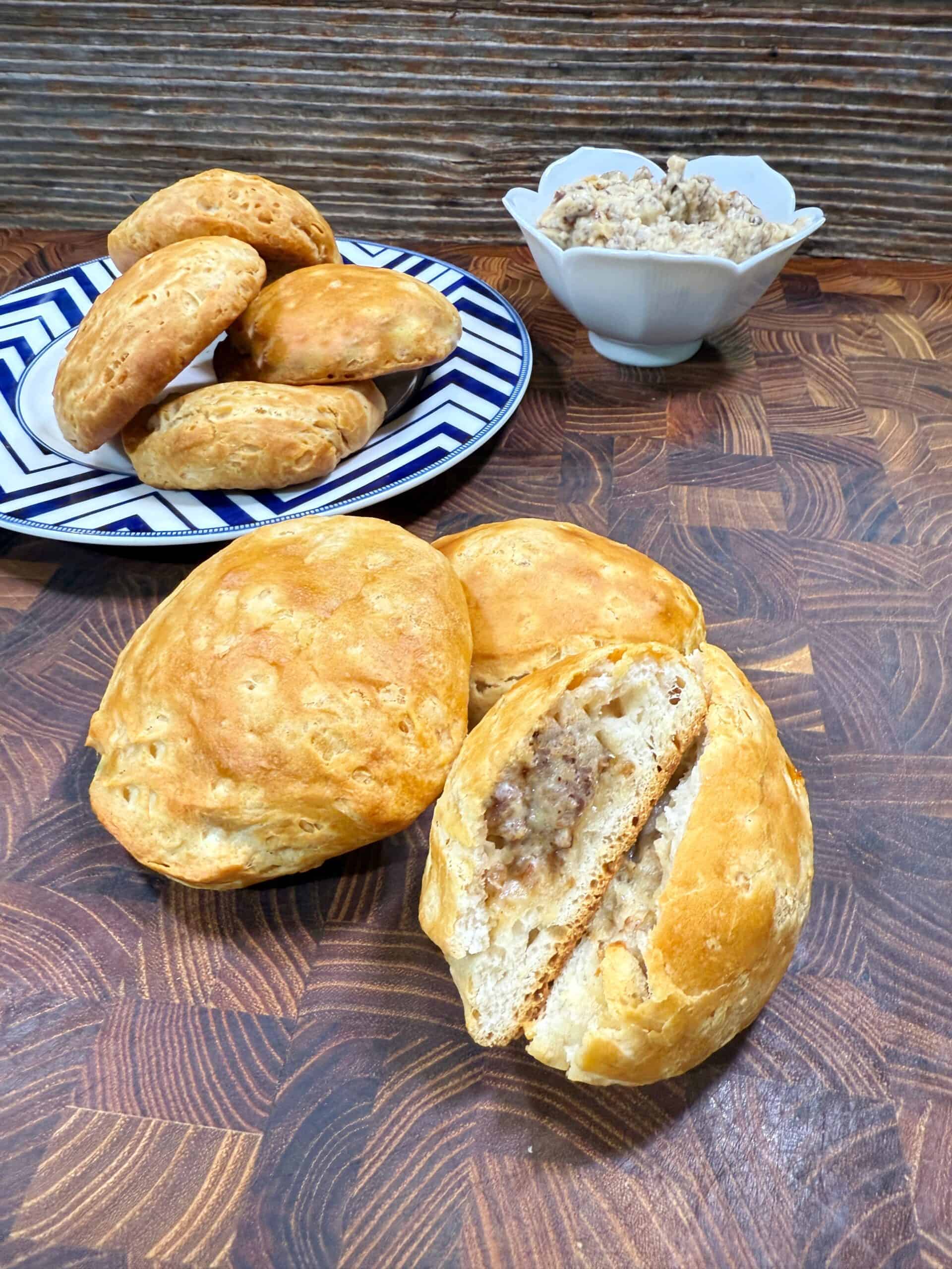 Golden brown bread rolls filled with sausage and cheese are displayed on a wooden surface. Some are on a blue and white plate, a few are in front, with one split open to show the filling. A bowl of sausage mixture sits in the background.