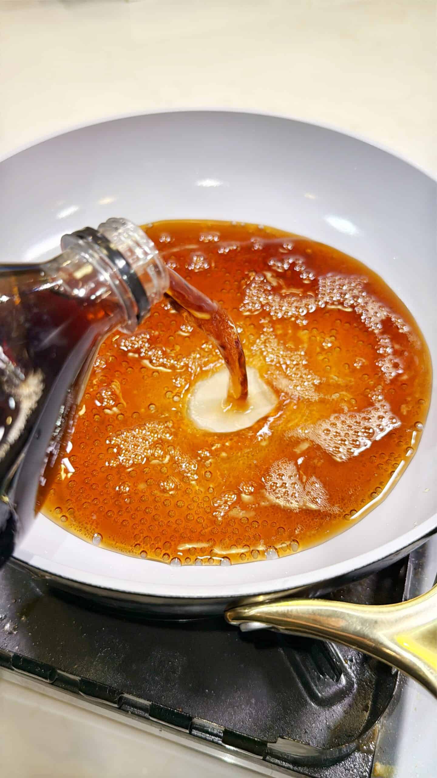 A close-up of a hand pouring dark soda from a bottle into a white frying pan on a stove, with bubbling liquid filling the pan.