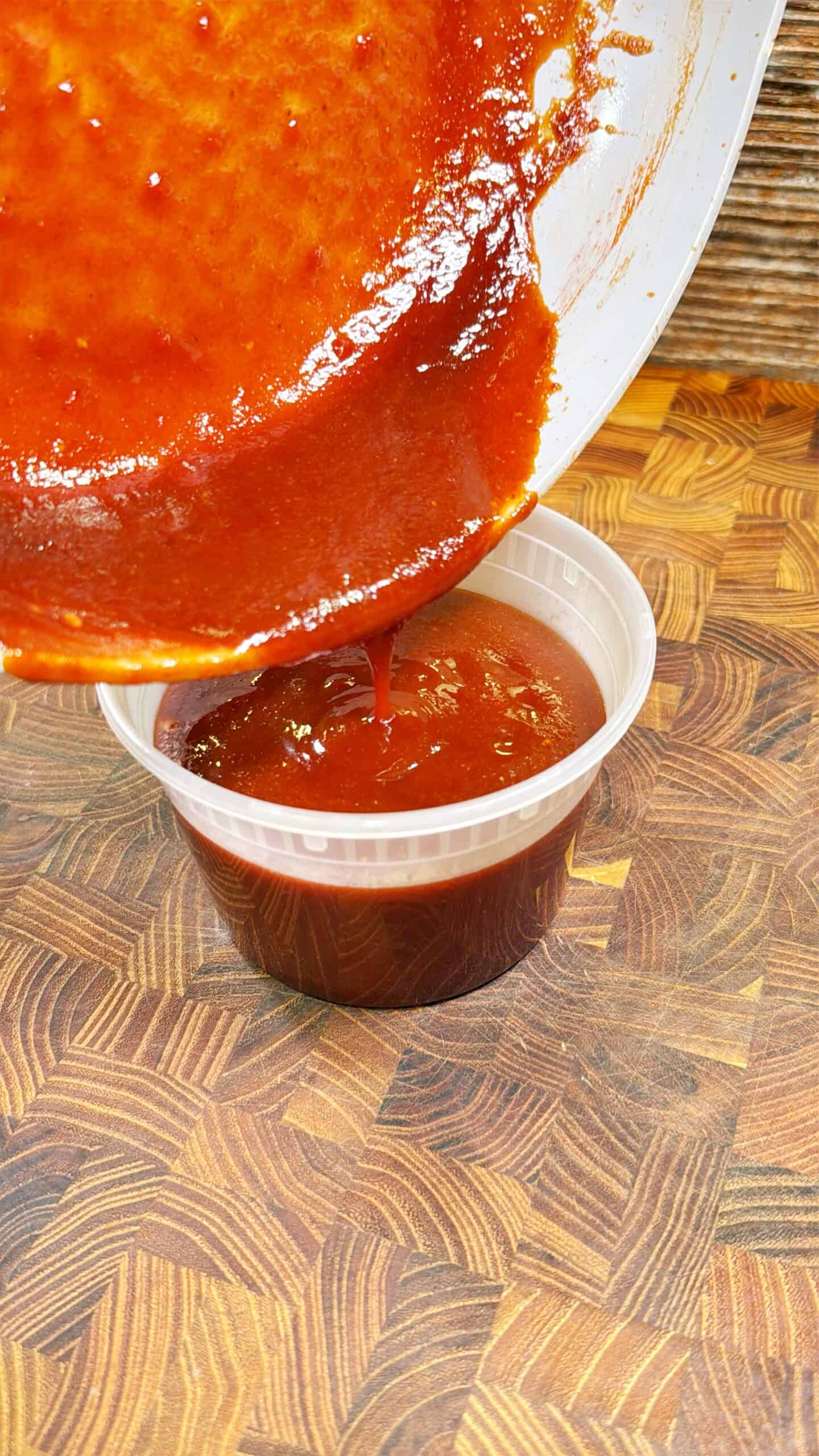 A thick, reddish-brown sauce is being poured from a white bowl into a small, clear plastic container on a patterned wooden surface.