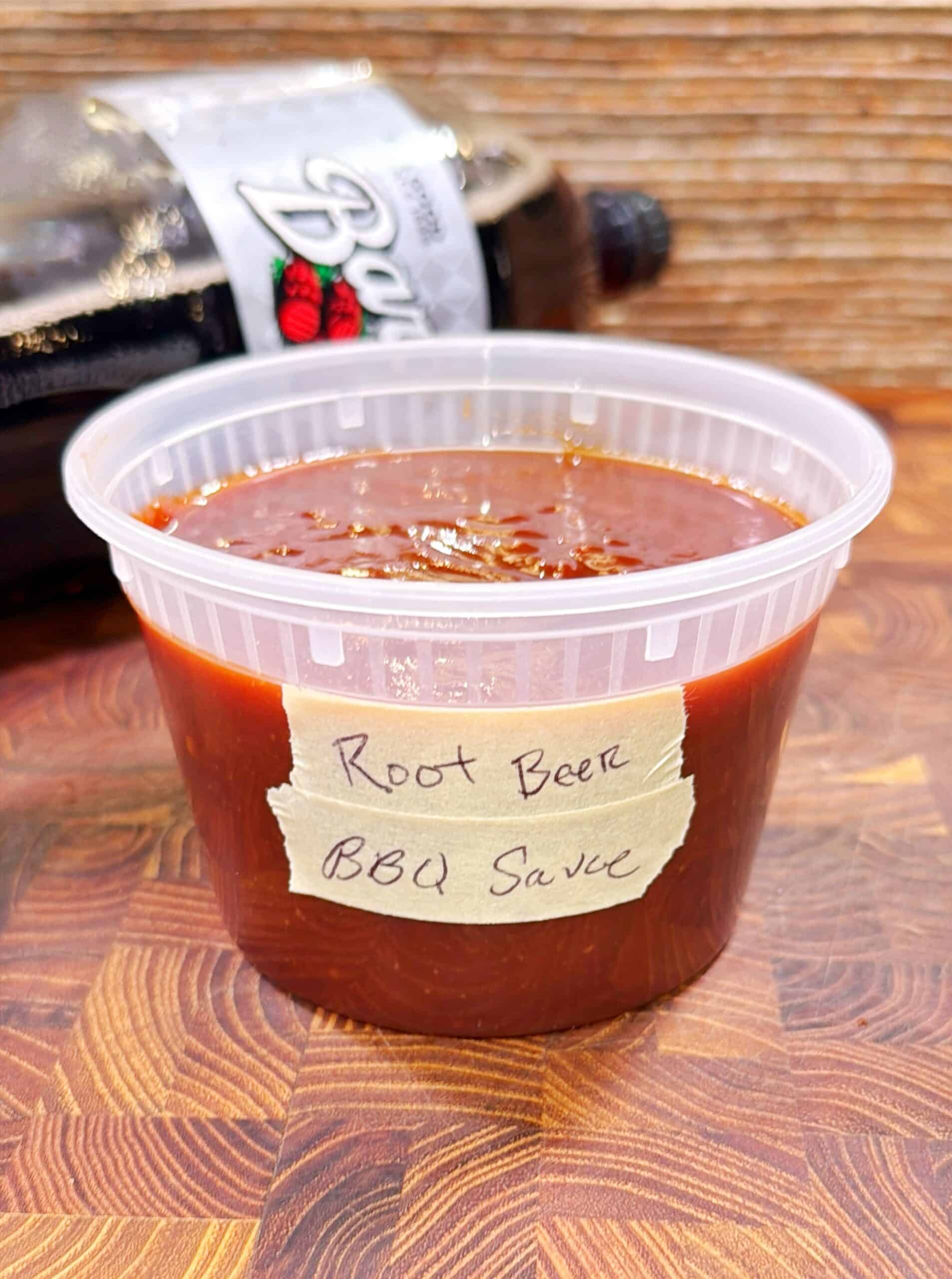A clear plastic container filled with dark brown BBQ sauce labeled Root Beer BBQ Sauce, sits on a wooden surface, with a bottle of root beer in the background.