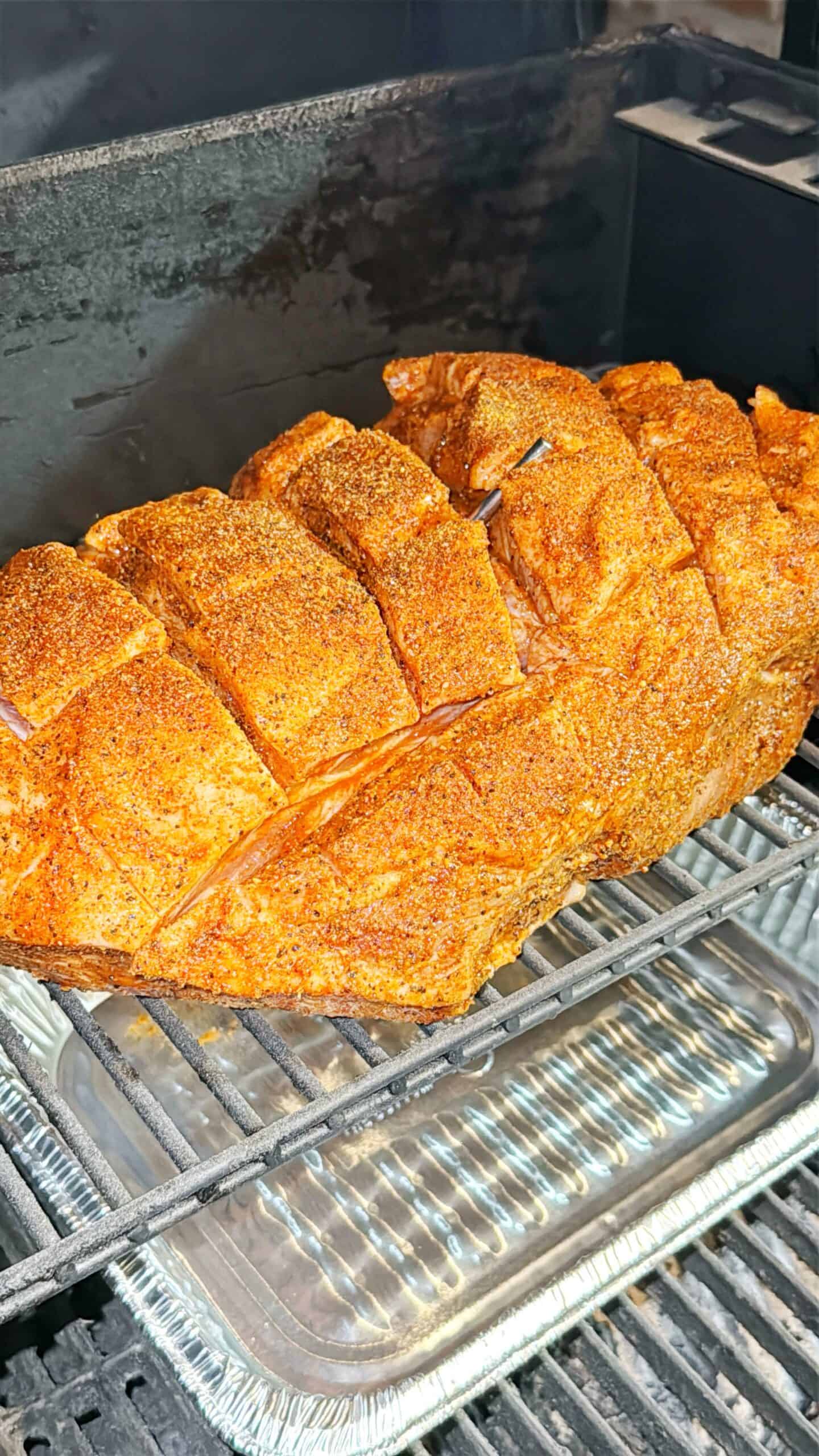 A large, seasoned pork roast with scored skin is cooking on a metal rack inside a smoker, with aluminum trays below to catch drippings. The surface of the meat is coated in spices.