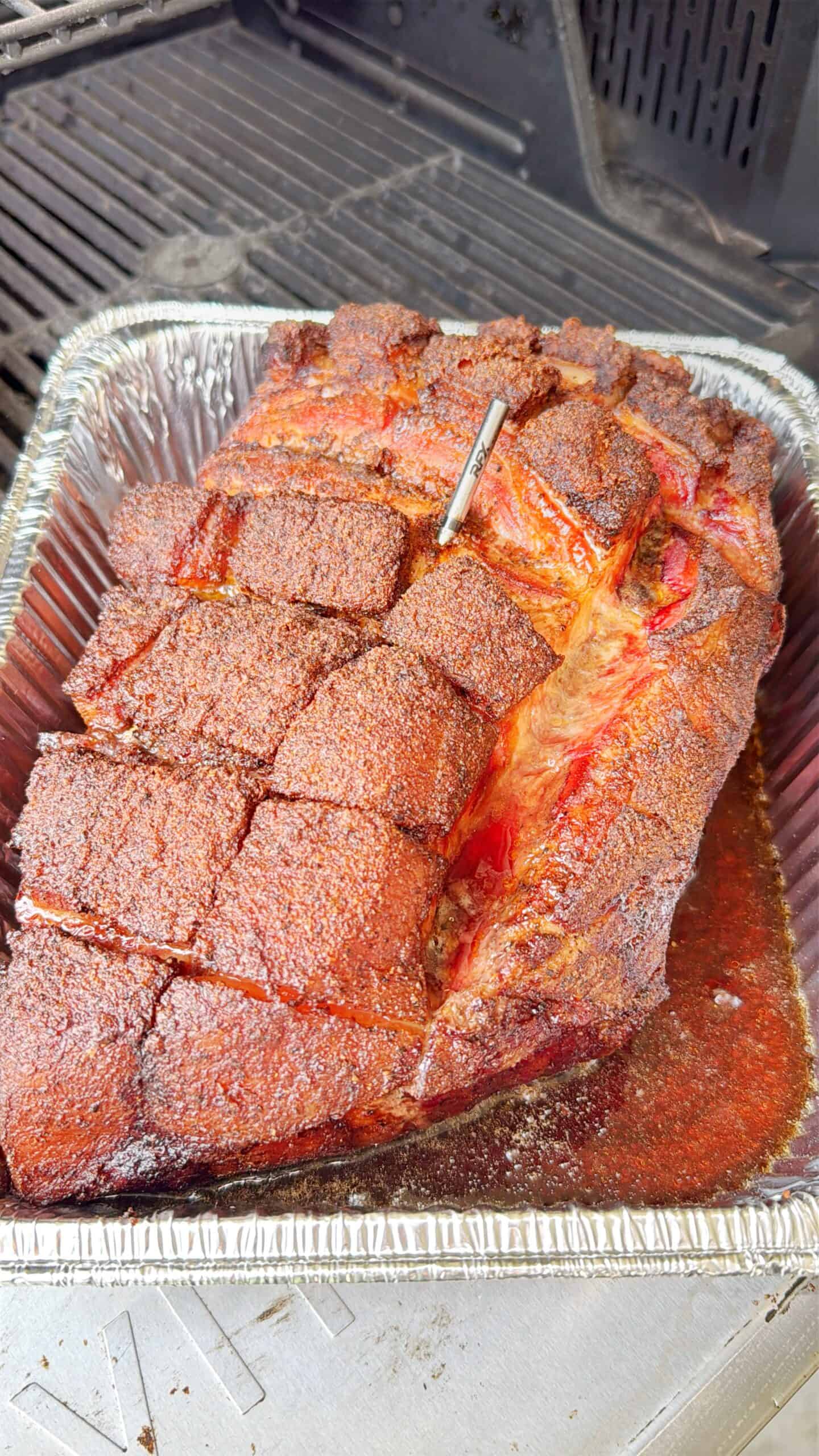 A large, seasoned beef brisket with a checkered cut sits in a foil pan on a grill. A meat thermometer is inserted into the brisket, and juices are pooled at the bottom of the pan.