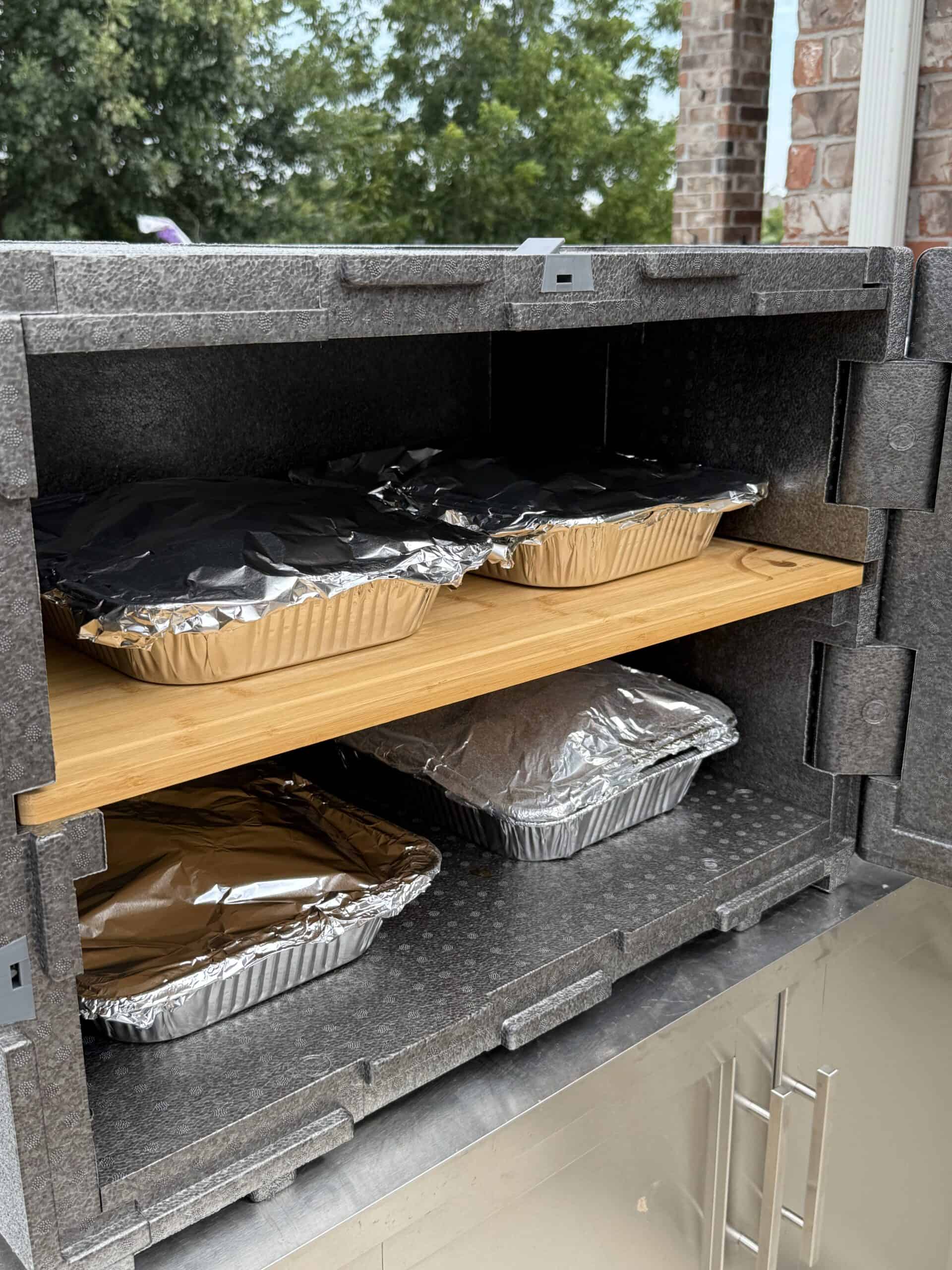 Four foil-covered aluminum trays with food are stored inside a gray insulated food carrier with a wooden shelf, situated outdoors near a brick wall and greenery.