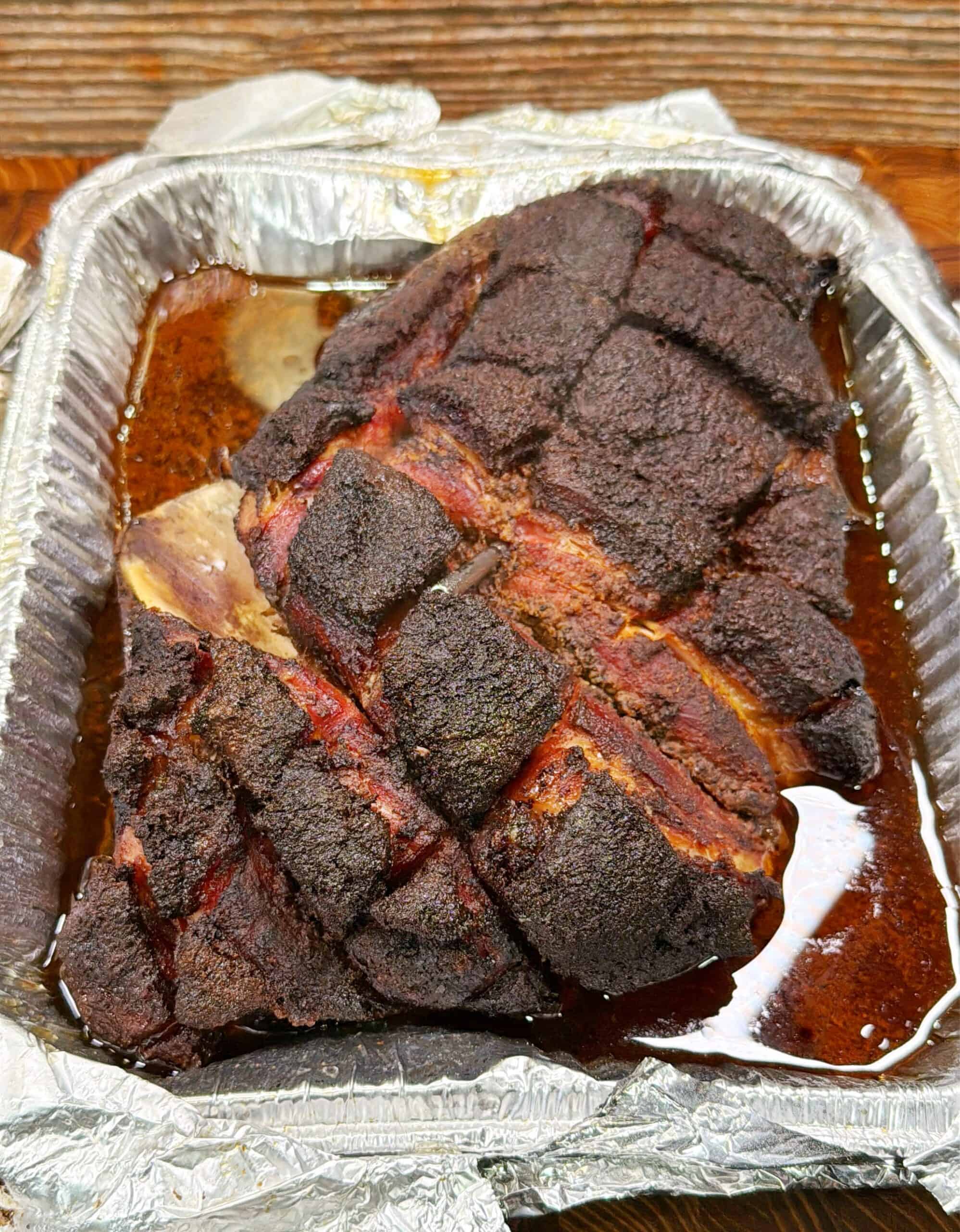 A cooked, seasoned pork roast with a dark, crispy crust sits in a disposable aluminum tray with juices pooling at the bottom. The meat is scored with a crosshatch pattern.