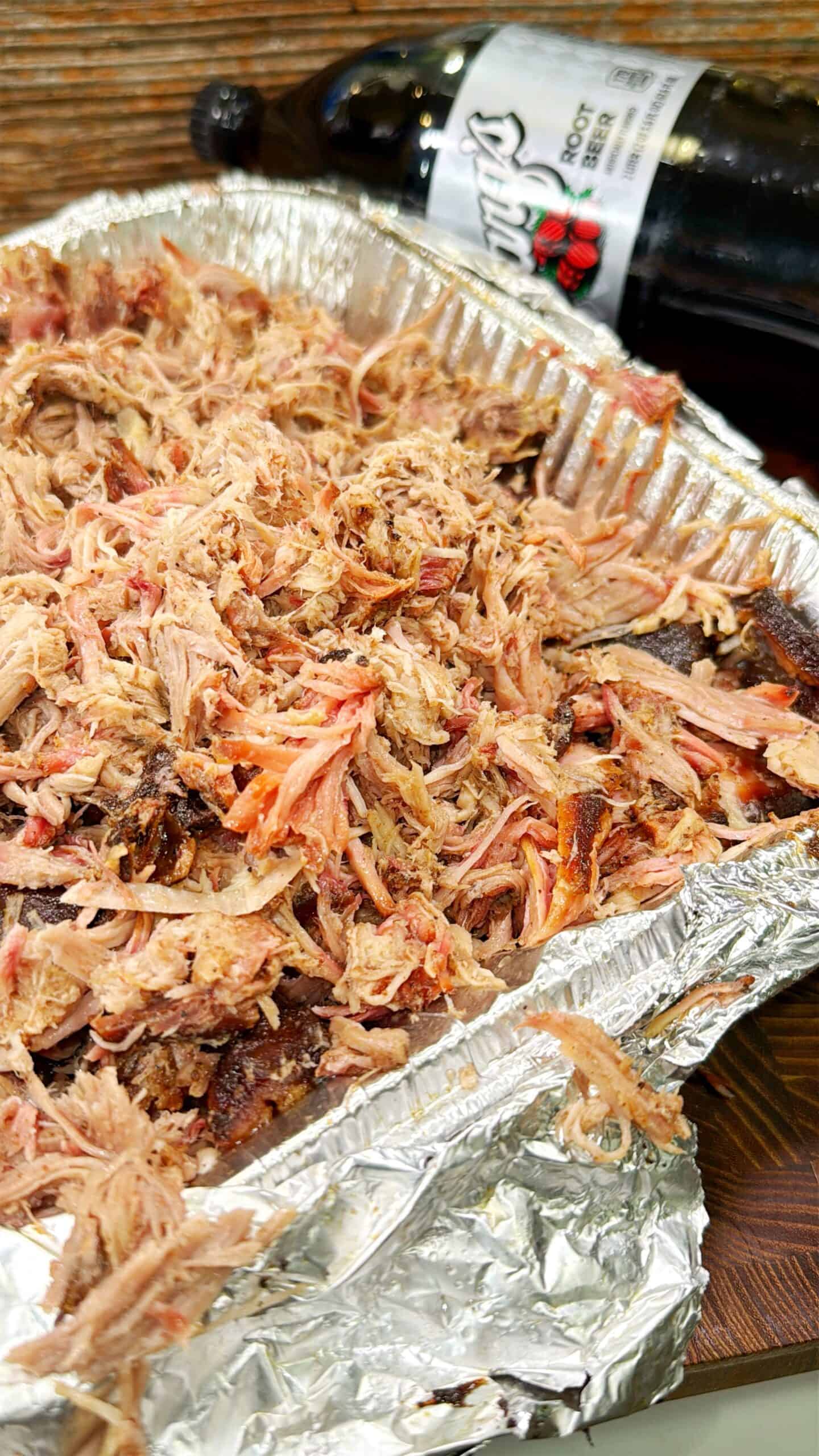 A close-up of shredded pulled pork in a disposable aluminum tray, with a bottle of root beer partially visible in the background.