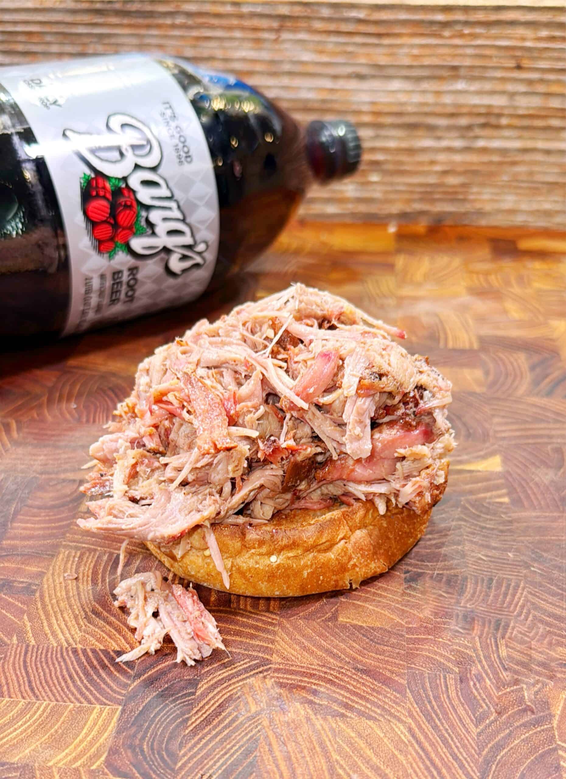 A pile of shredded barbecue pork sits on a toasted bun, placed on a wooden cutting board. In the background, a bottle of Barq’s root beer lies on its side.