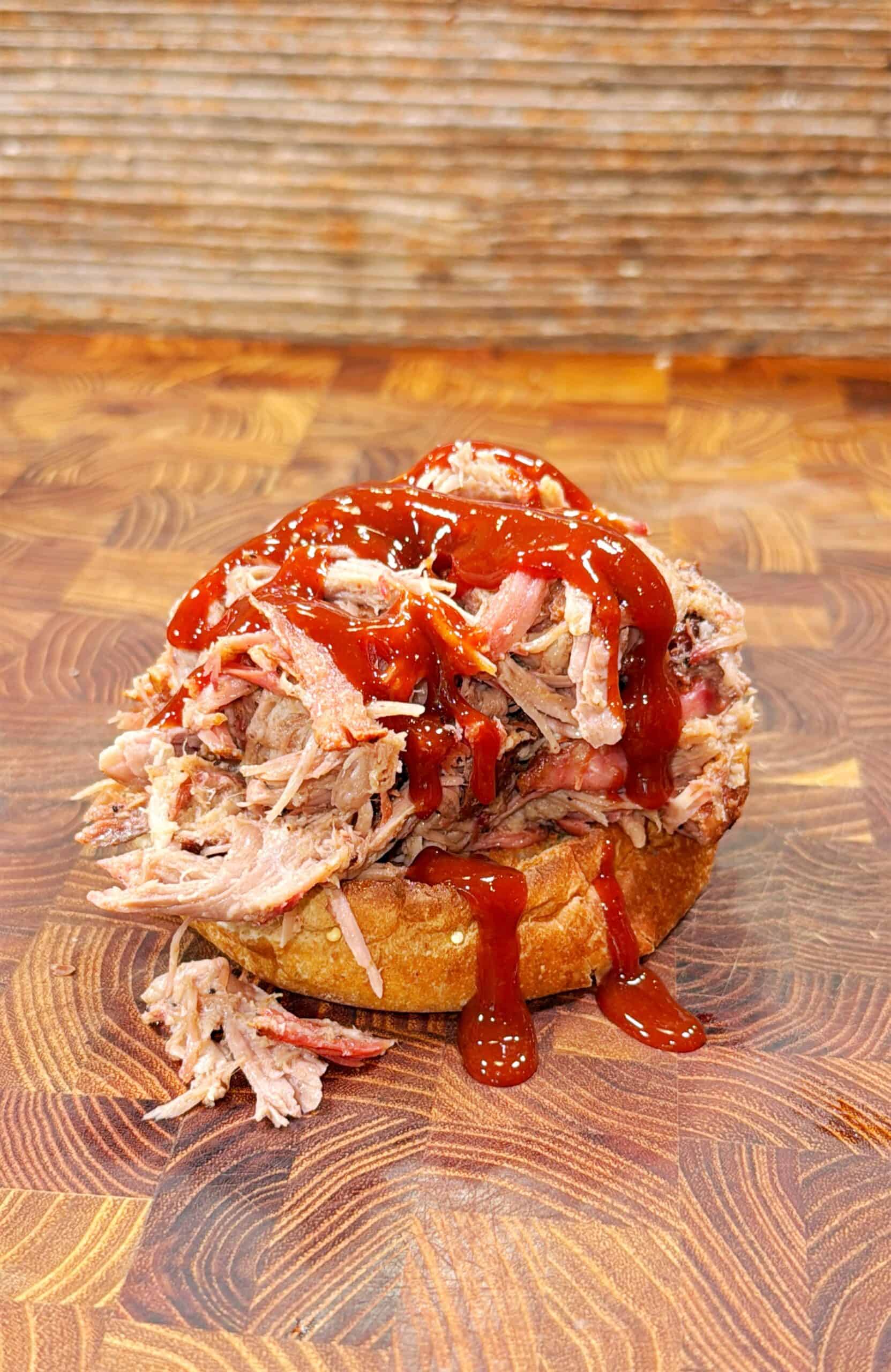 A pulled pork sandwich topped with barbecue sauce sits on a wooden surface, with shredded meat spilling over the sides of a toasted bun.