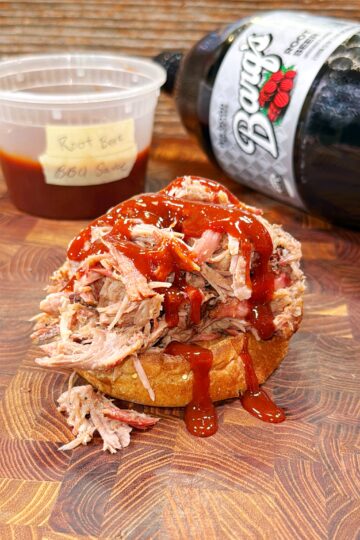 A pulled pork sandwich on a wooden surface, topped with barbecue sauce. Behind it is a plastic container labeled Root Beer BBQ Sauce and a bottle of root beer lying on its side.