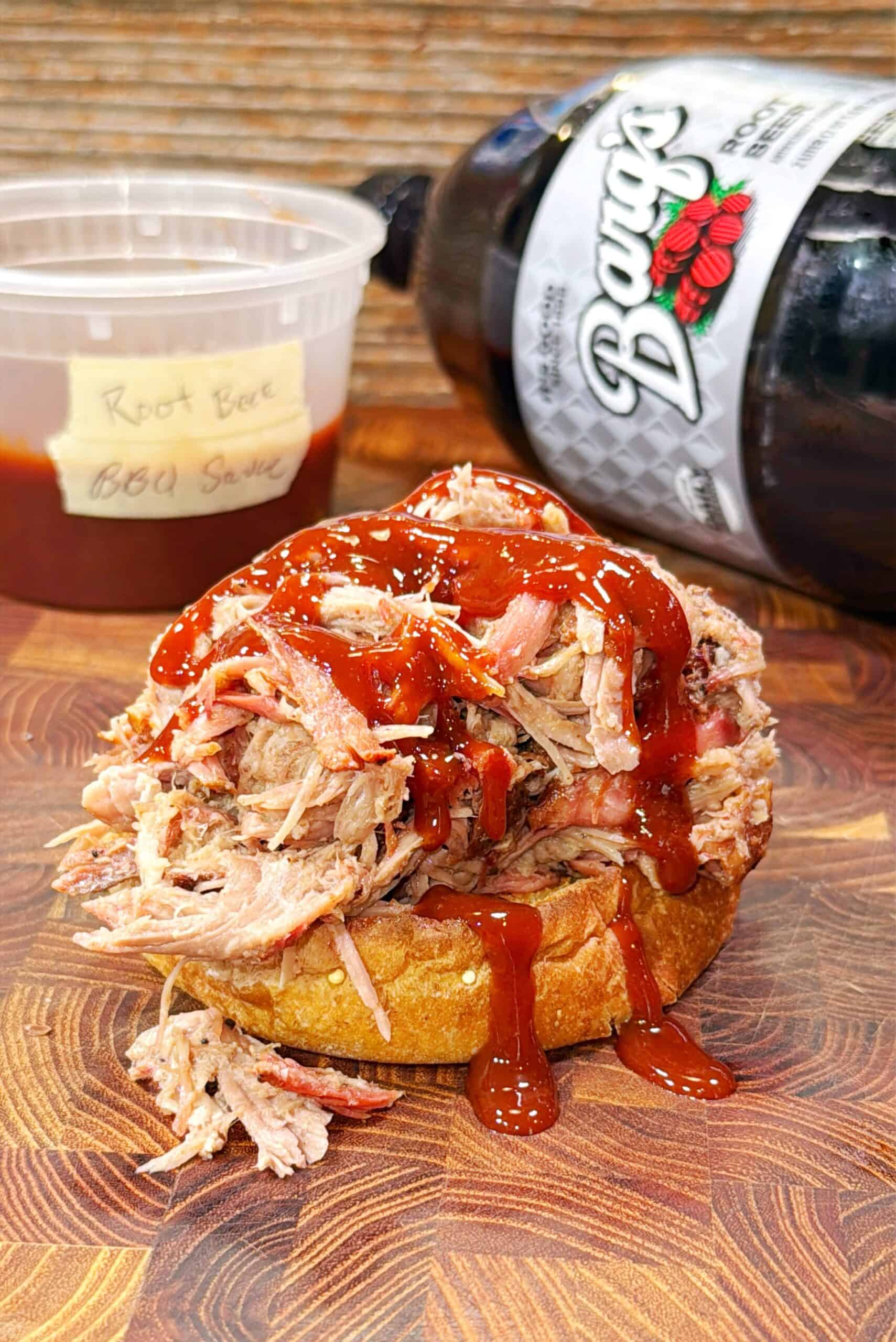A root beer pulled pork sandwich topped with BBQ sauce sits on a wooden surface. Behind it are a plastic container labeled Root Beer BBQ Sauce and a tilted bottle of root beer.