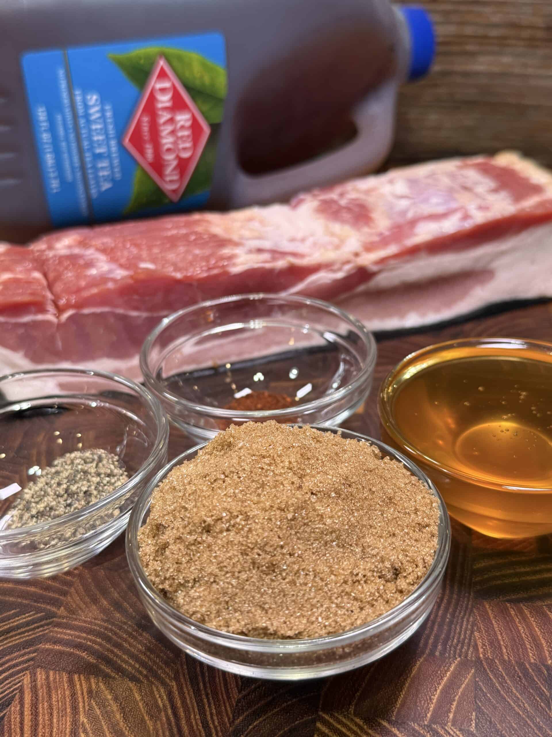 Several ingredients on a wooden surface, including a slab of raw pork, a bottle of sweet tea, bowls of black pepper, brown sugar, and honey.