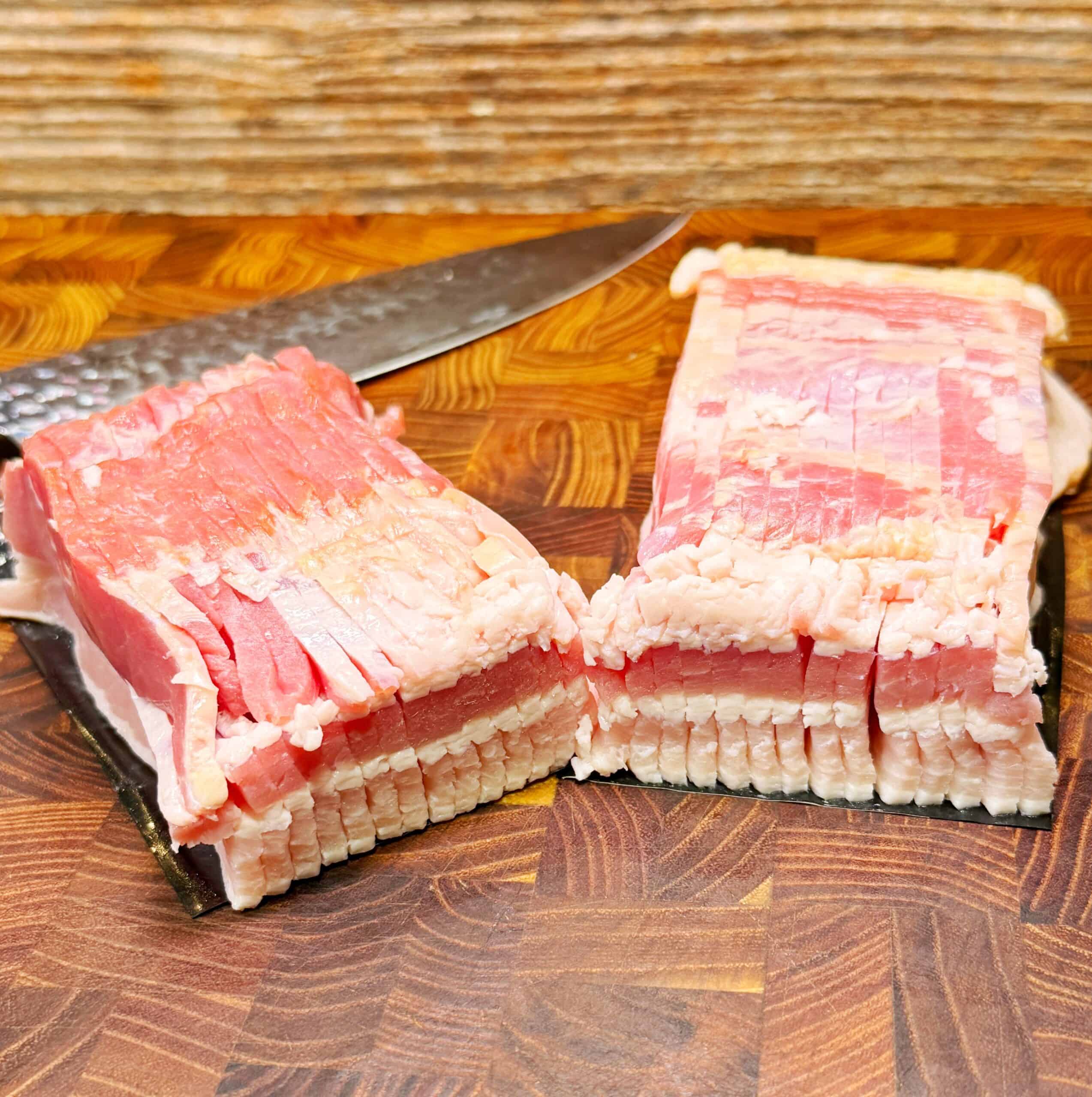 Two neatly stacked blocks of uncooked bacon slices sit on a patterned wooden cutting board with a large knife placed behind them. The bacon is pink with visible layers of fat.