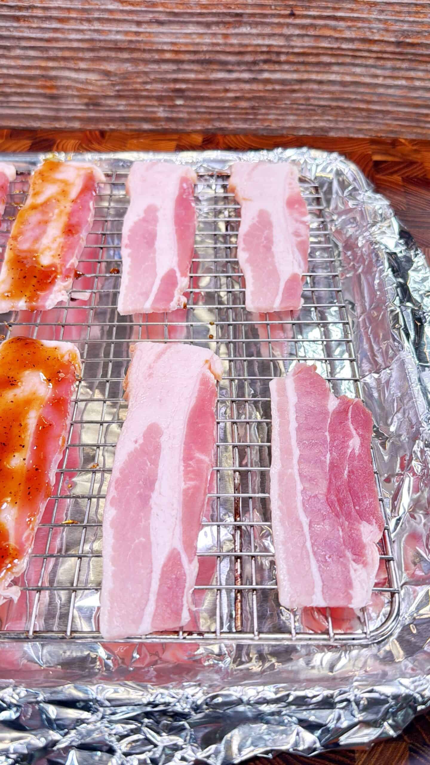 Raw bacon strips are arranged on a wire rack set over a foil-lined baking sheet. One strip on the left is coated with a brown sauce, while the others remain plain and uncooked. The background is a wooden surface.