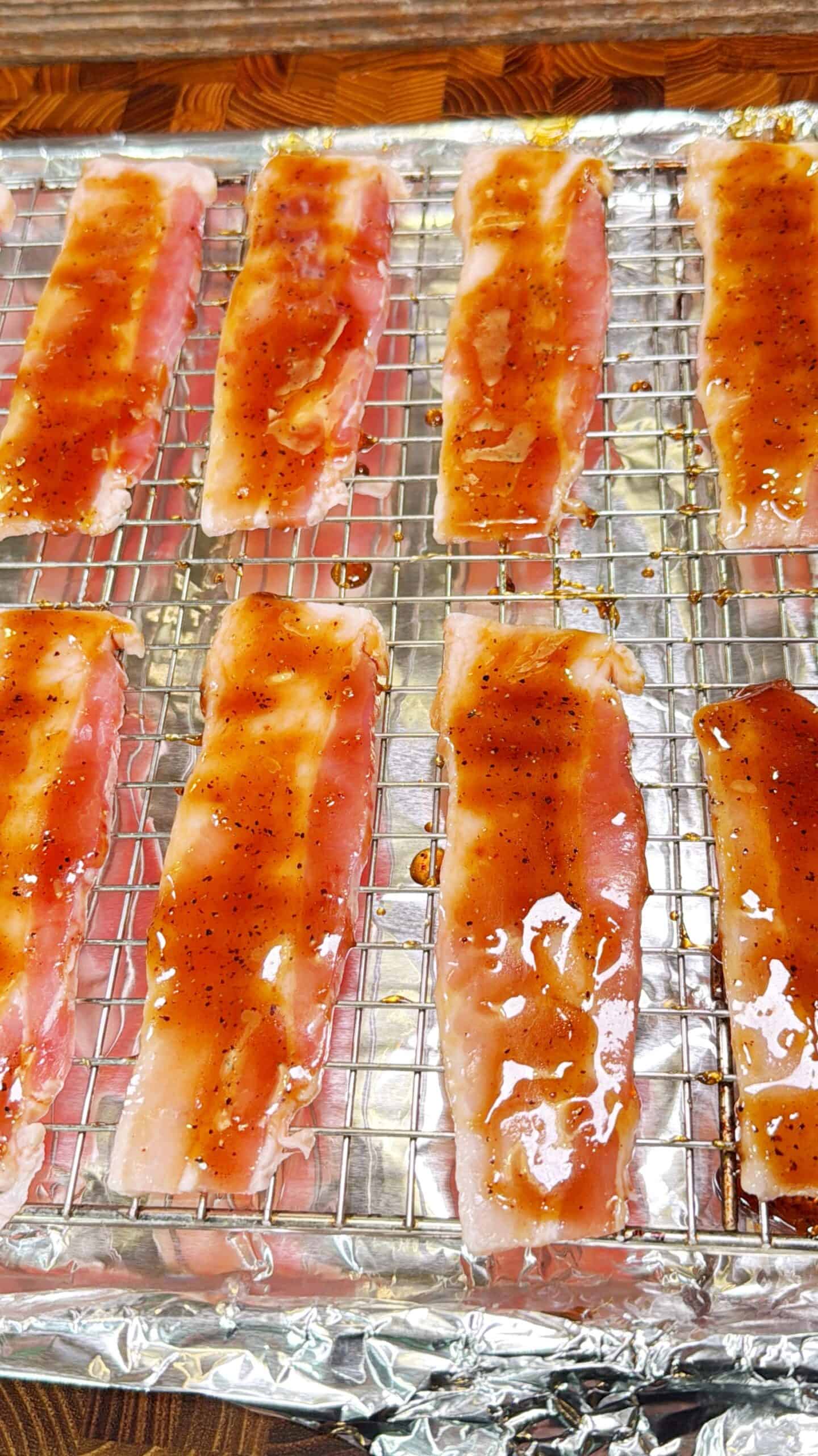 Strips of raw bacon coated with a glossy sauce are arranged on a wire rack over a foil-lined baking sheet, ready to be cooked.