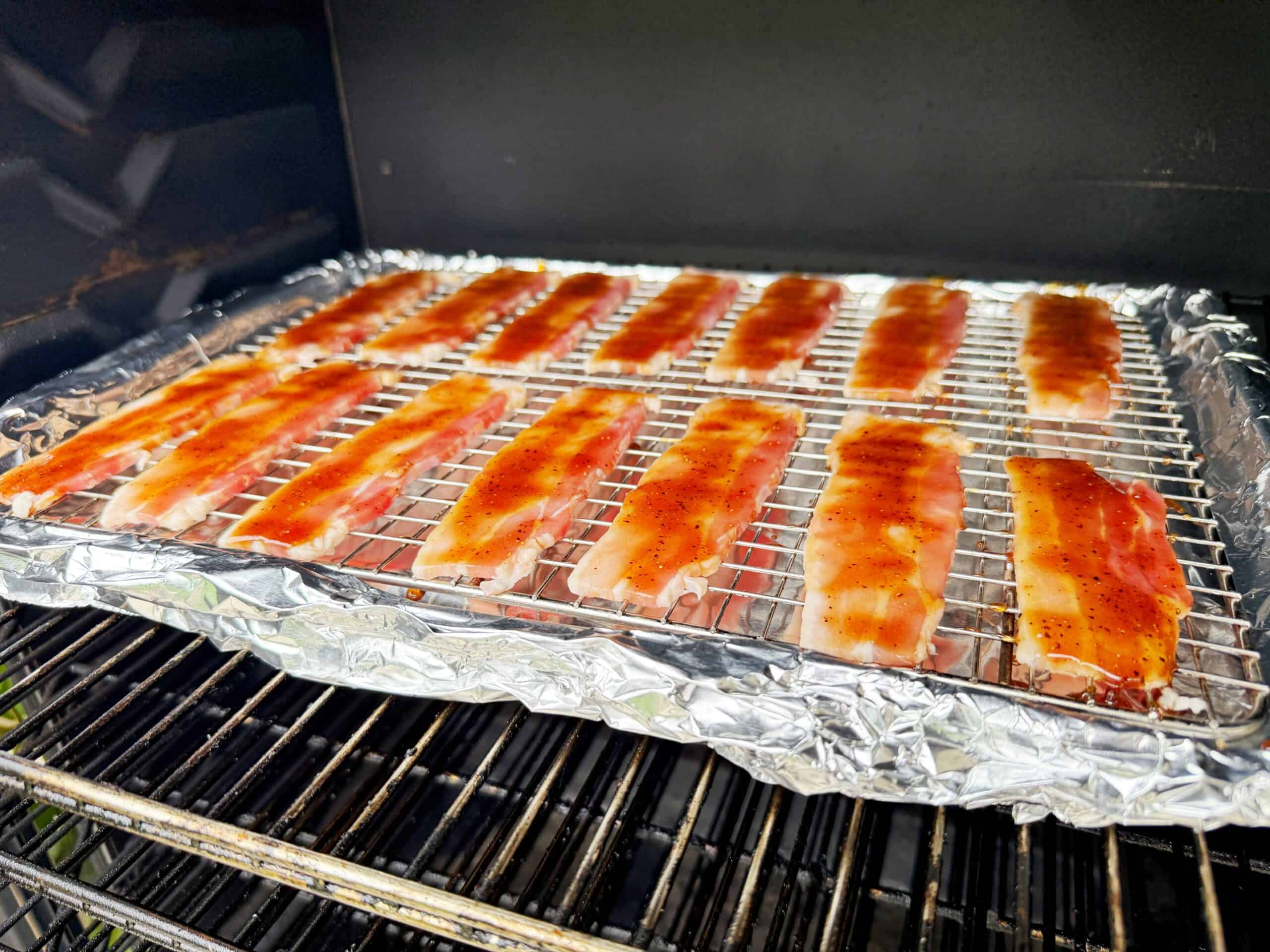 Uncooked bacon slices coated with sauce are arranged on a wire rack over a foil-lined tray inside an oven or smoker.