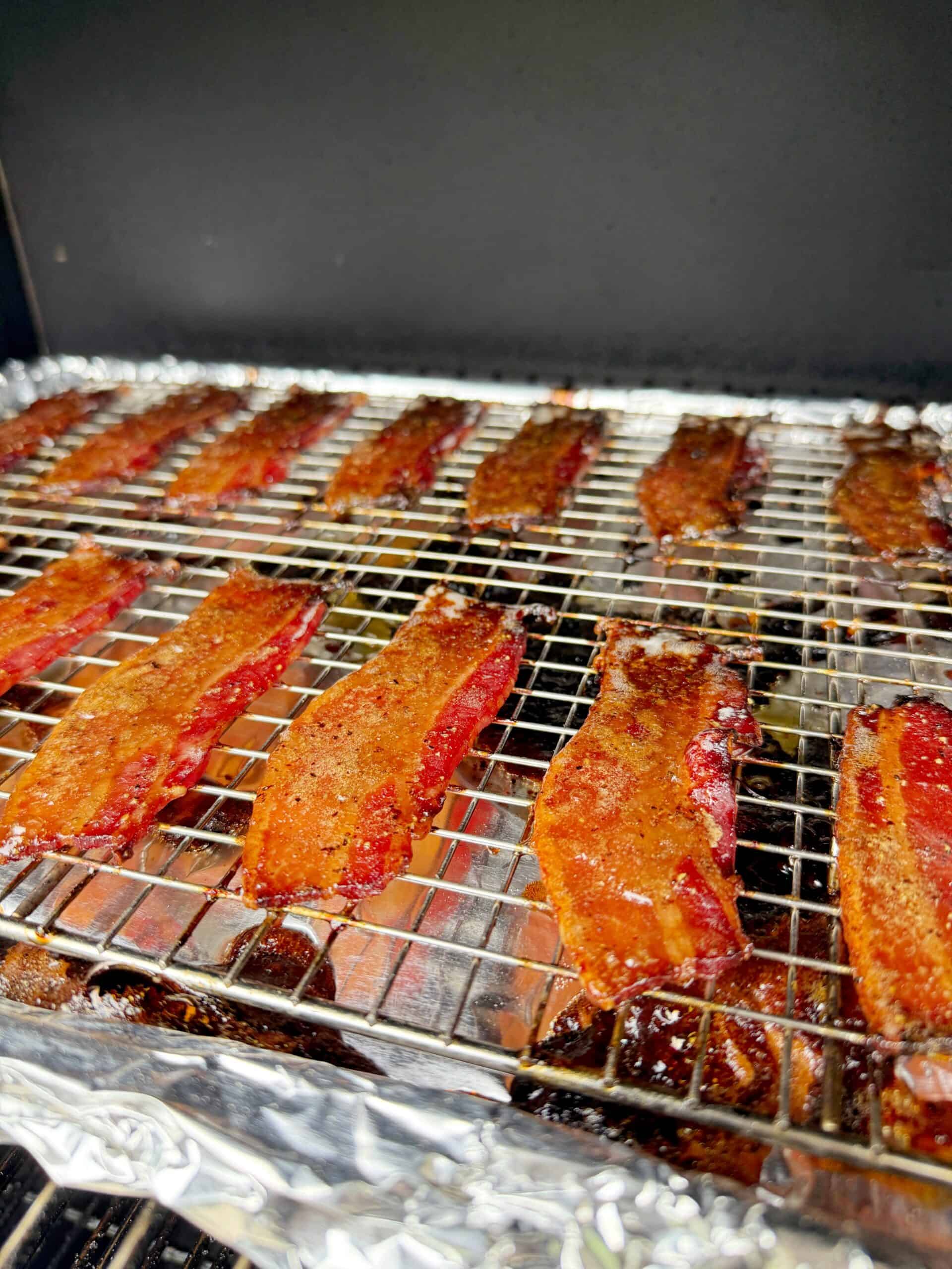 Rows of thick, glazed bacon strips are arranged on a wire rack over a foil-lined baking sheet, being cooked in an oven or smoker. The bacon appears crispy and caramelized.
