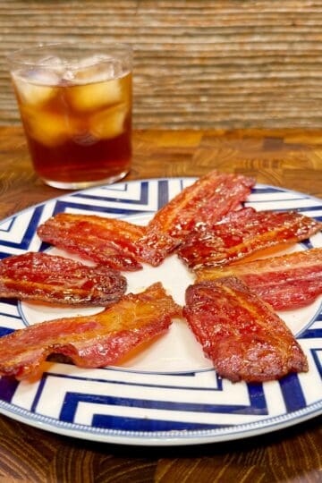 A plate with several strips of crispy glazed bacon arranged in a circle, placed on a geometric-patterned plate, with a glass of iced dark beverage in the background.