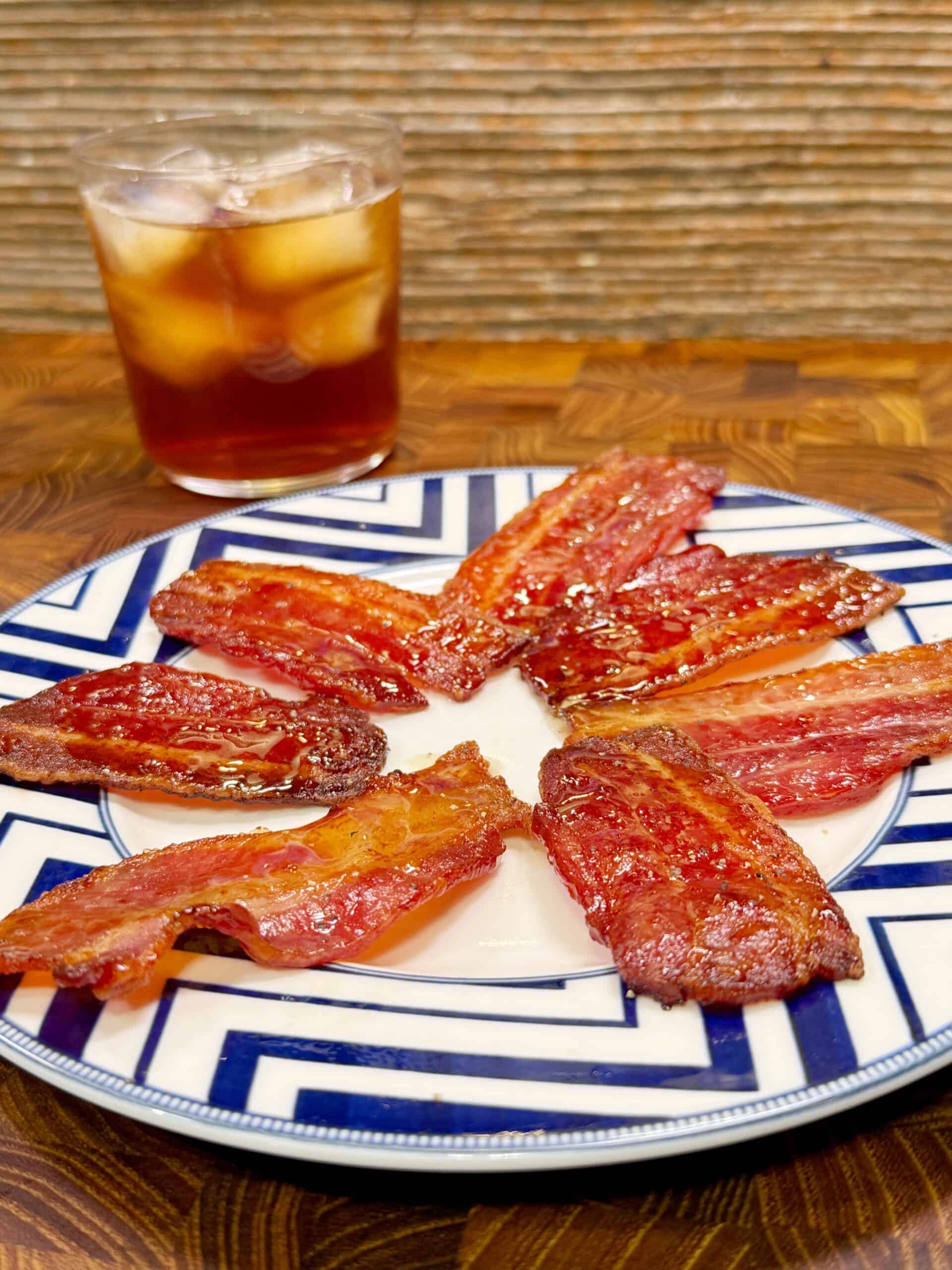 A plate with several strips of crispy glazed sweet tea candied bacon arranged in a circle, placed on a geometric-patterned plate, with a glass of iced dark beverage in the background.