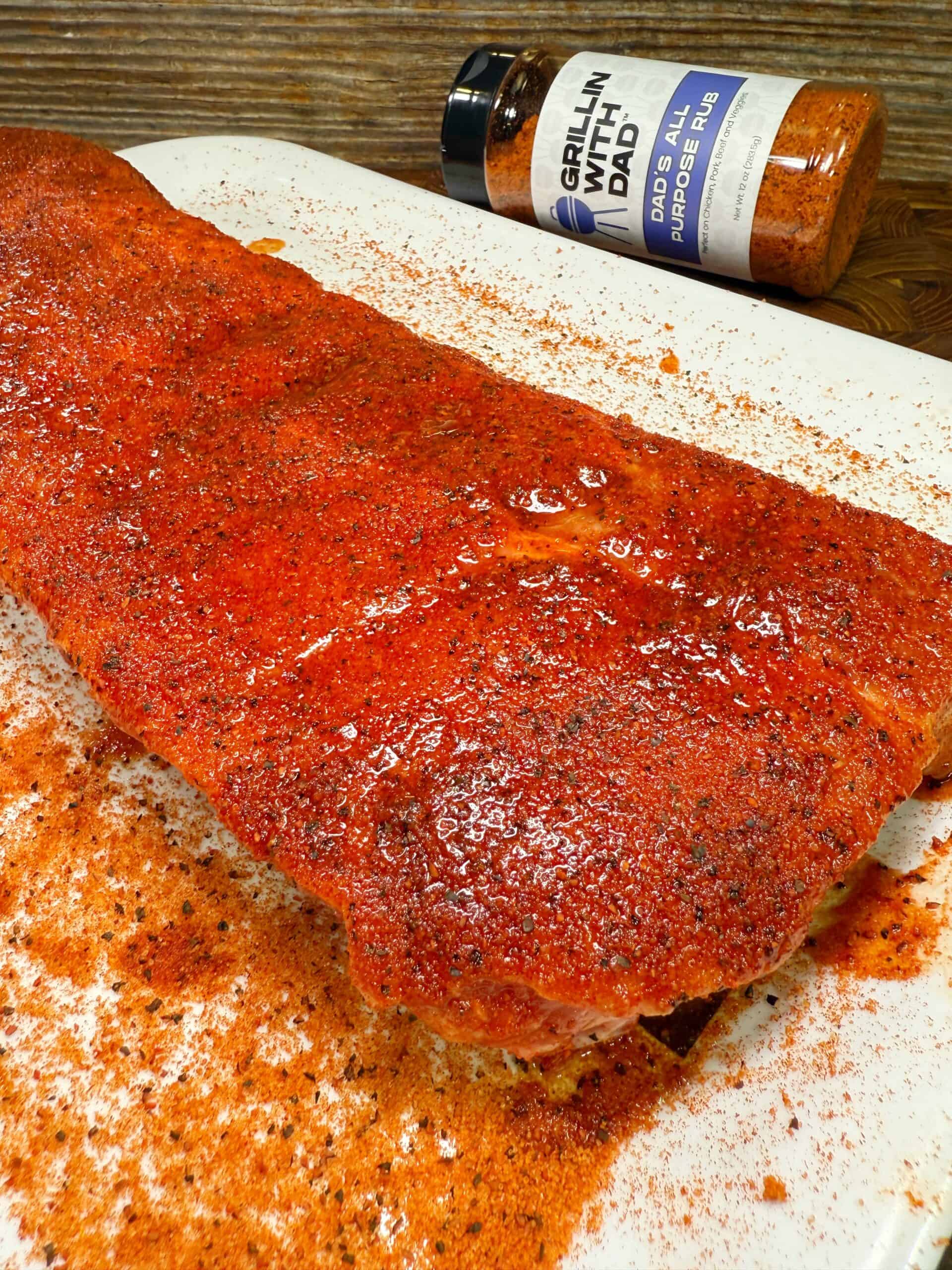 A slab of raw ribs coated with a reddish spice rub sits on a white tray. In the background, a bottle of Grillin With Dad All Purpose Rub seasoning is visible on a wooden surface.