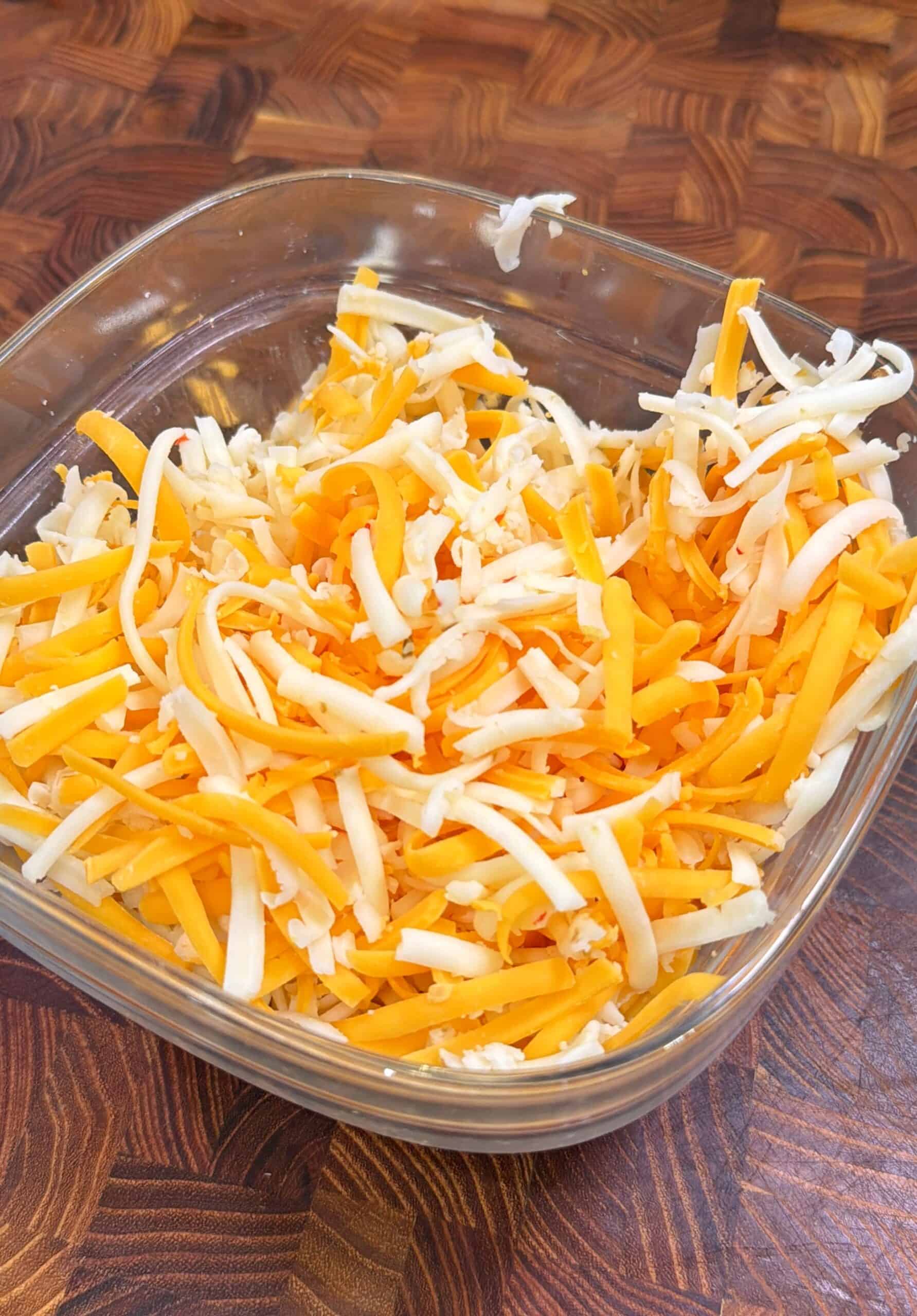A glass bowl filled with shredded yellow and white cheese sits on a wooden surface with a geometric pattern.