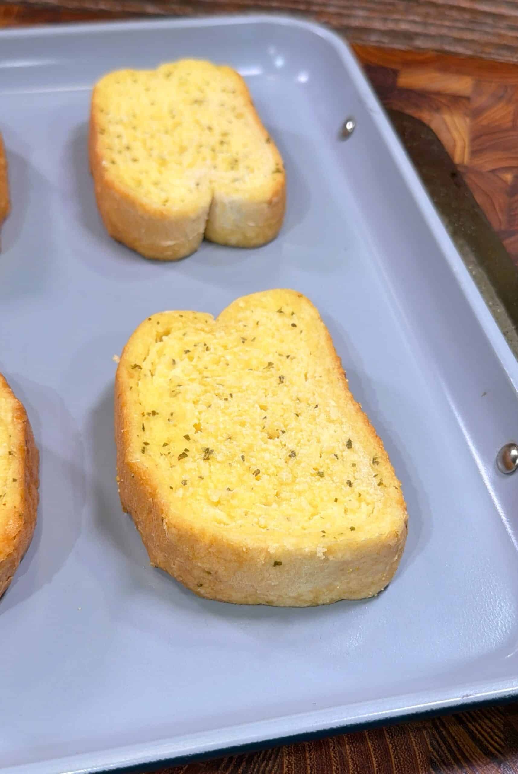 A close-up of two slices of garlic bread on a light gray baking sheet, resting on a wooden surface. The bread appears golden and seasoned with herbs.