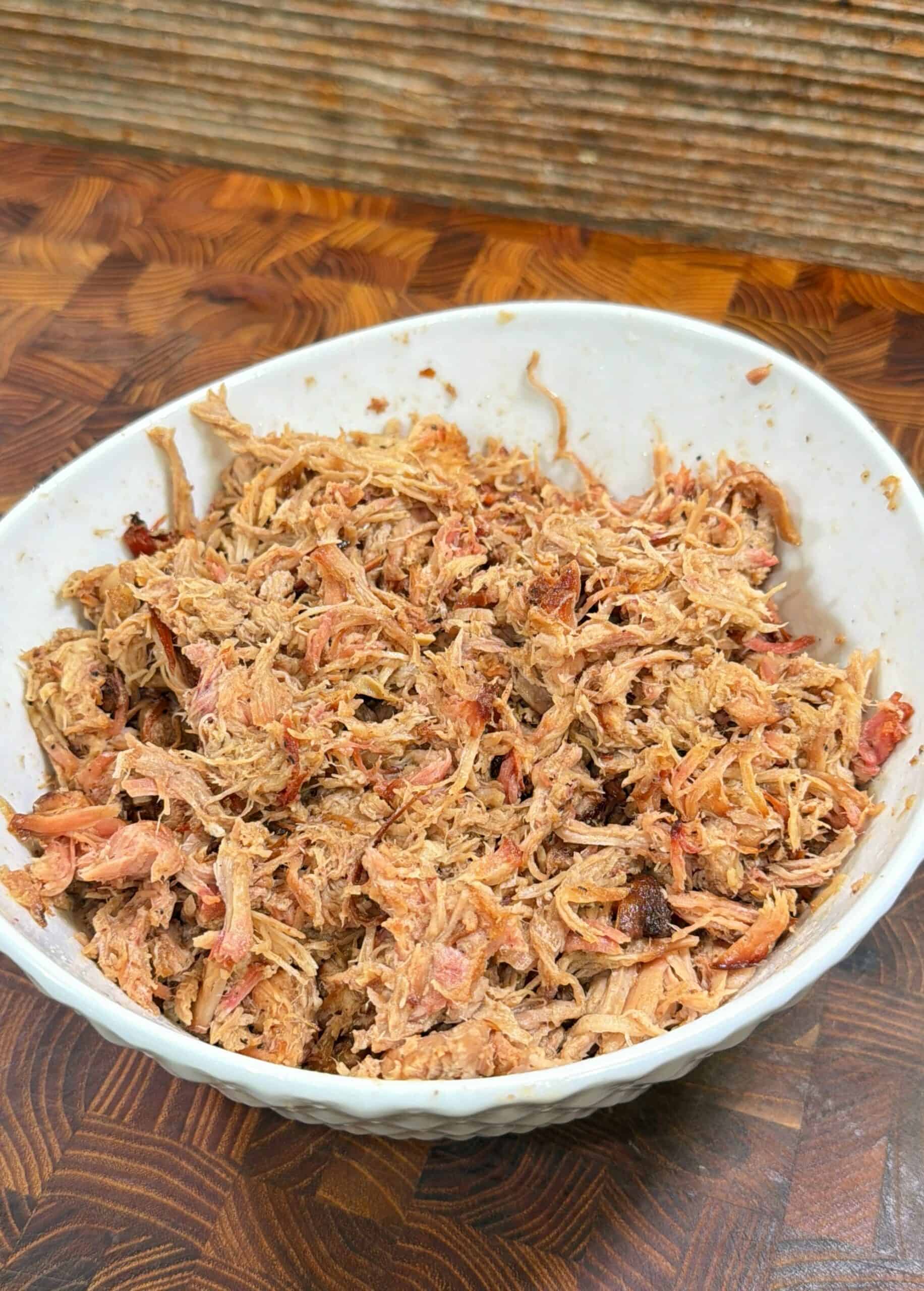 A white bowl filled with shredded pulled pork sits on a wooden surface, with a rustic, textured background behind it.