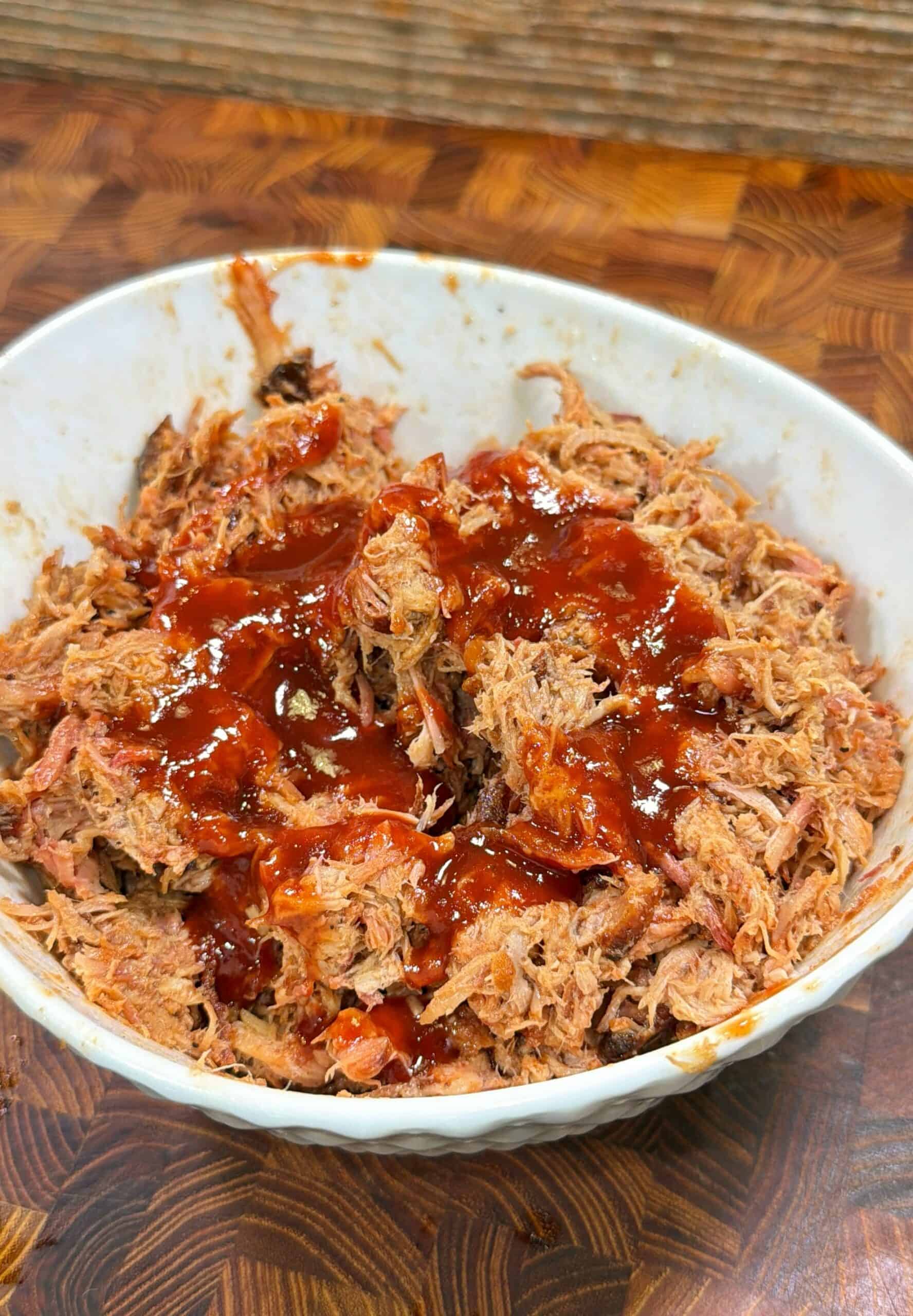 A white bowl filled with shredded pulled pork topped with a generous layer of barbecue sauce, placed on a brown patterned wooden surface.