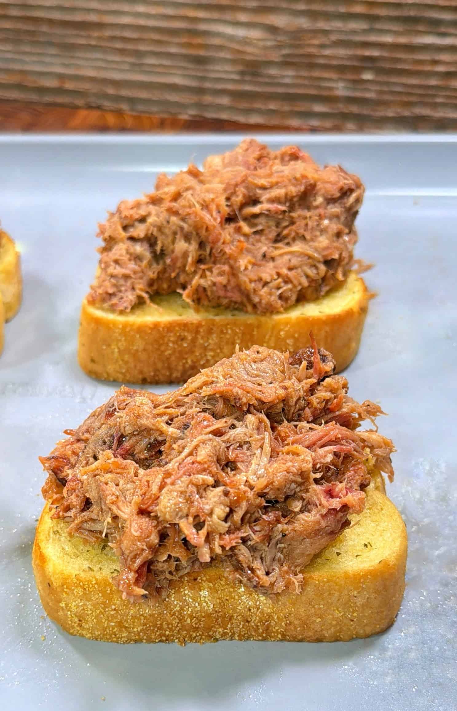 Sheet pan grilled cheese slices of toasted bread each topped with a generous serving of shredded pulled pork, placed on a gray surface with a wooden background.