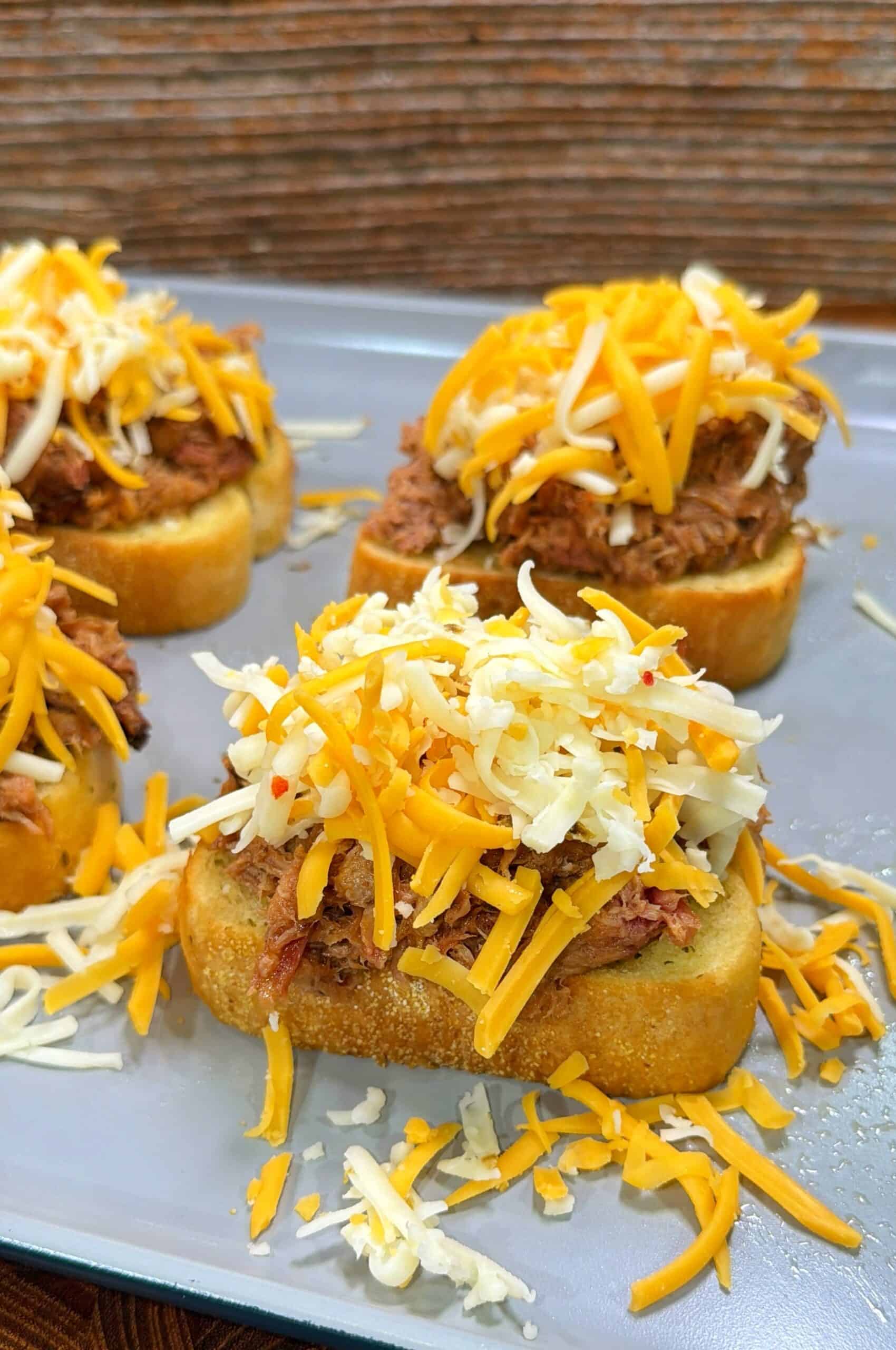 Three pieces of toasted bread topped with shredded meat and a generous amount of mixed shredded yellow and white cheese, arranged on a metal tray.