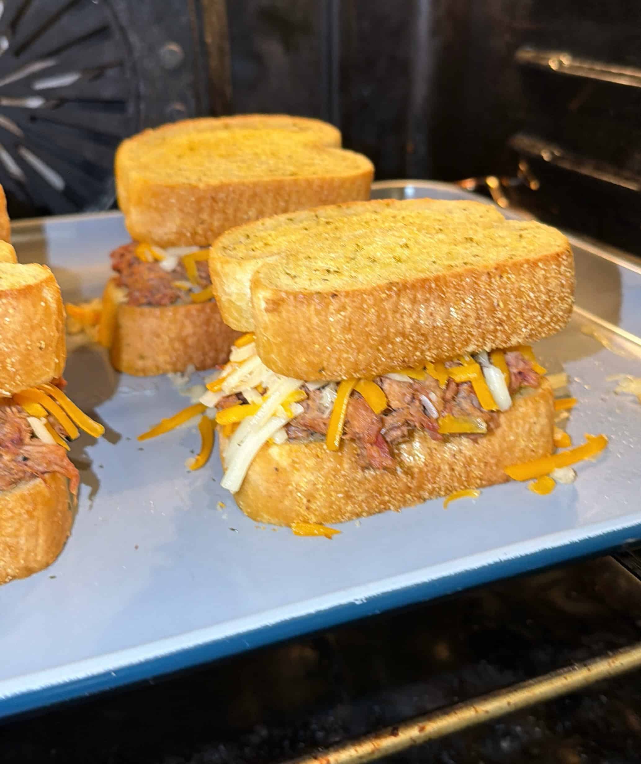 Three sandwiches with thick toasted bread, shredded cheese, and pulled meat are arranged on a baking tray inside an oven, ready to be baked or melted.