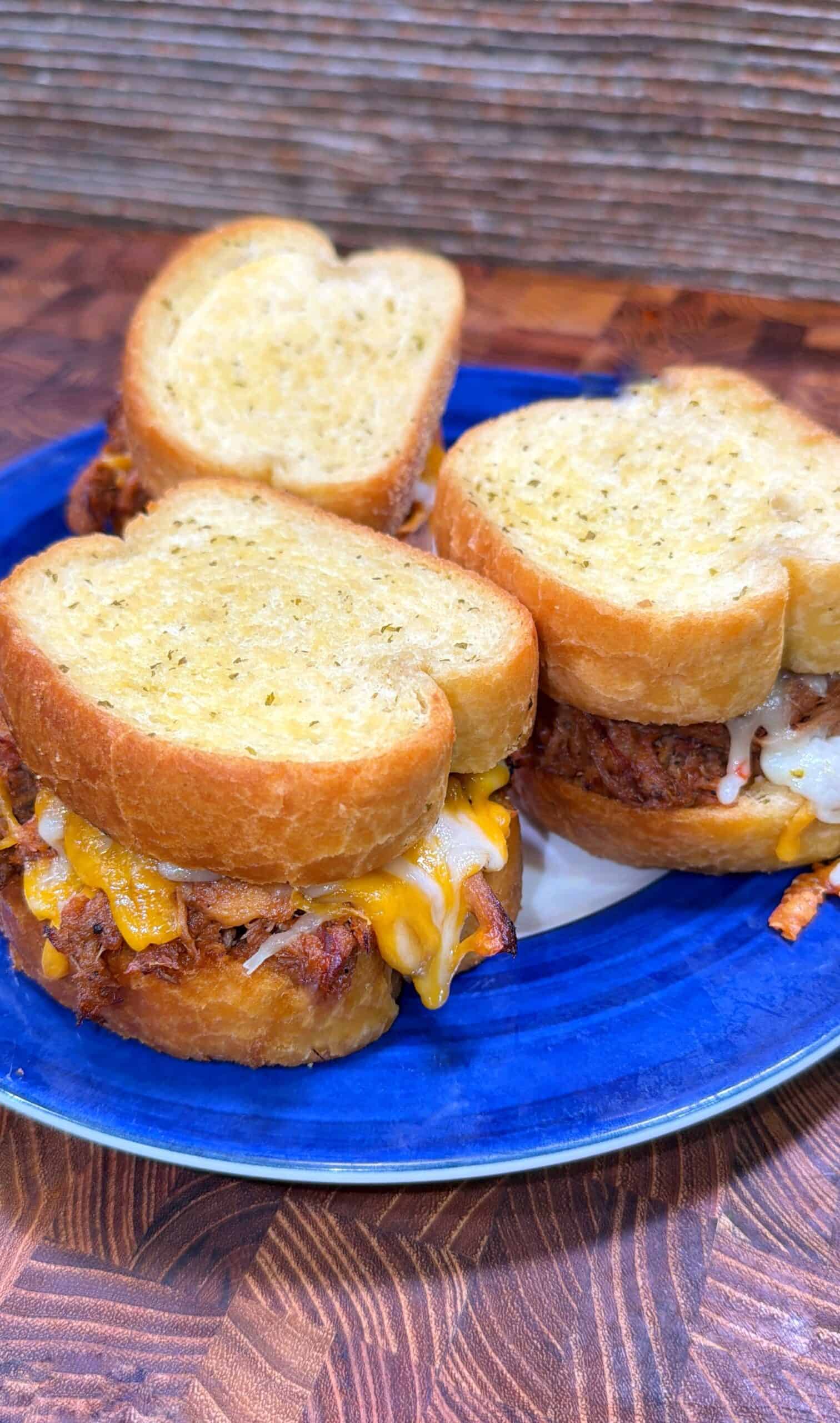 Three sandwiches with melted cheese and shredded meat filling, served on thick slices of toasted bread, are arranged on a blue plate atop a wooden surface.