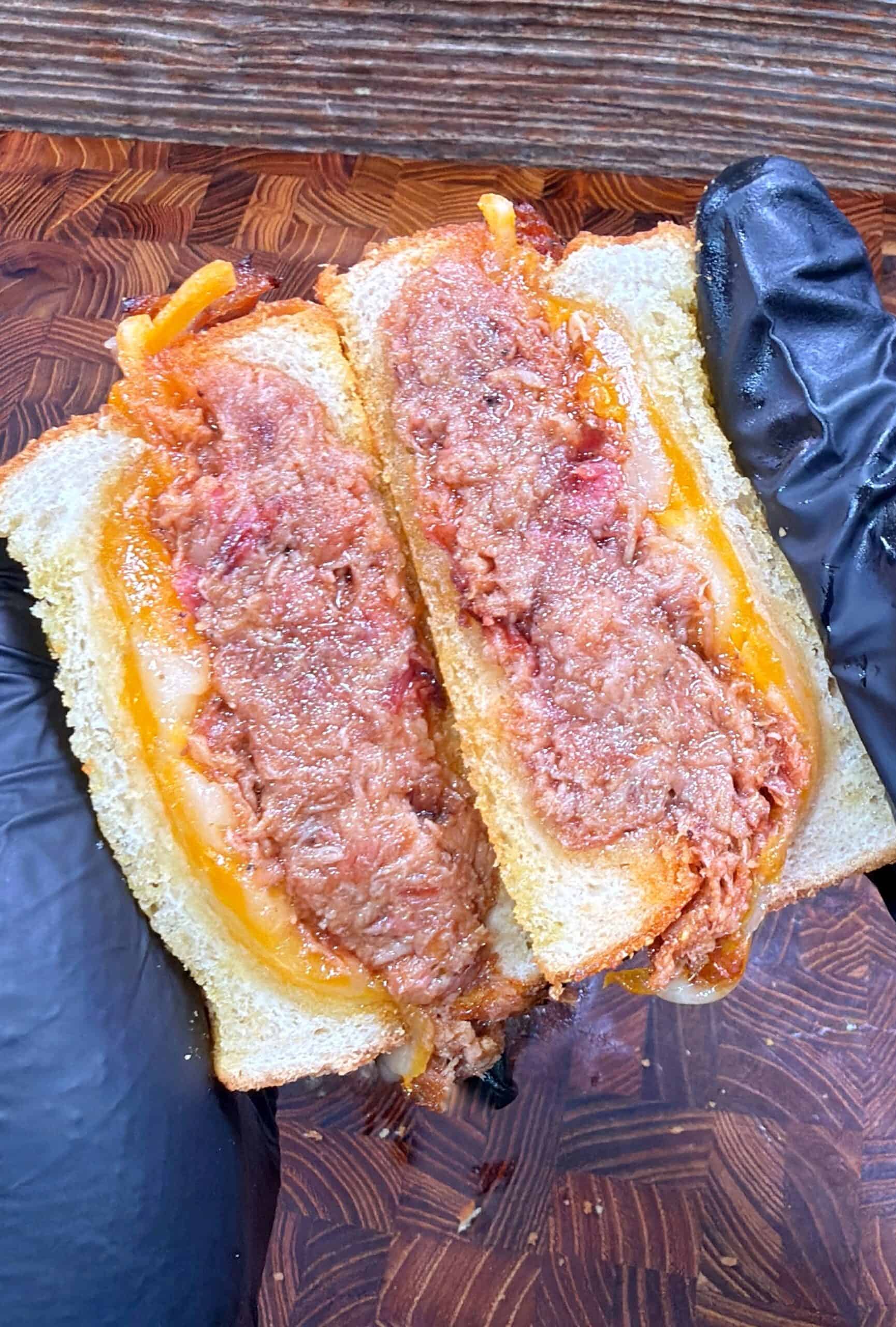 A gloved hand holds a sandwich on sliced white bread, filled with shredded meat and melted cheese, cut in half on a wooden cutting board.