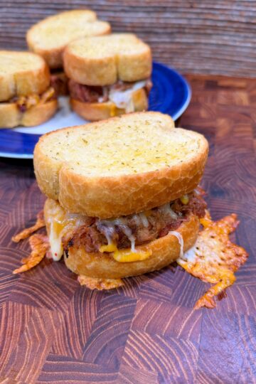 A cheesy, golden-brown slider sandwich with crispy edges sits on a wooden surface, with two more similar sandwiches on a blue plate in the background.