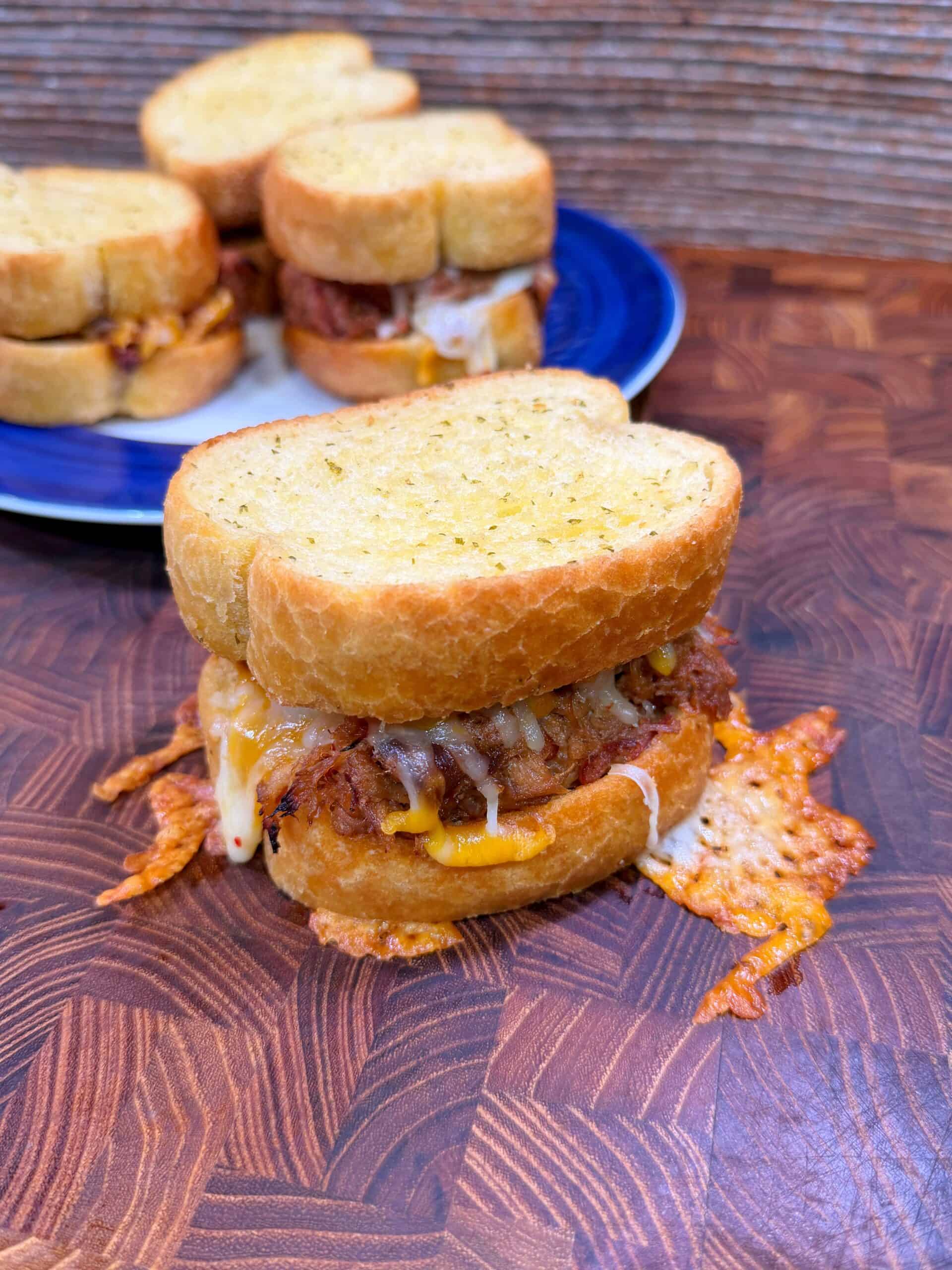 A cheesy, golden-brown slider sandwich with crispy edges sits on a wooden surface, with two more similar sandwiches on a blue plate in the background.