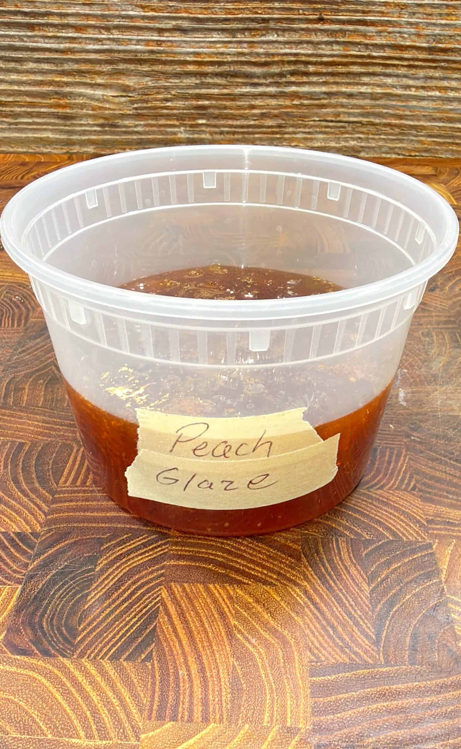 A clear plastic container filled with peach-colored glaze sauce sits on a wooden surface. A piece of masking tape labeled Peach Glaze is attached to the front, perfect for brushing over peach glazed ribs.