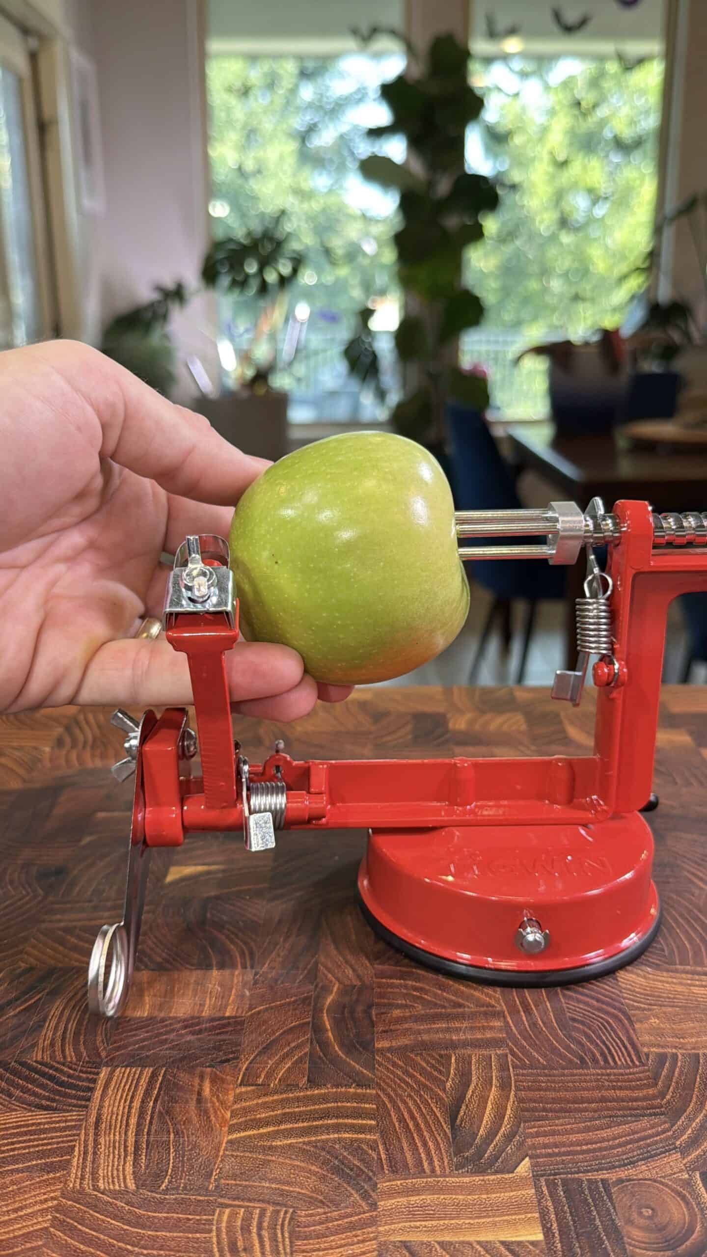 A hand places a green apple onto a red apple peeler and corer machine on a wooden countertop, with a bright room and green plants visible in the background.
