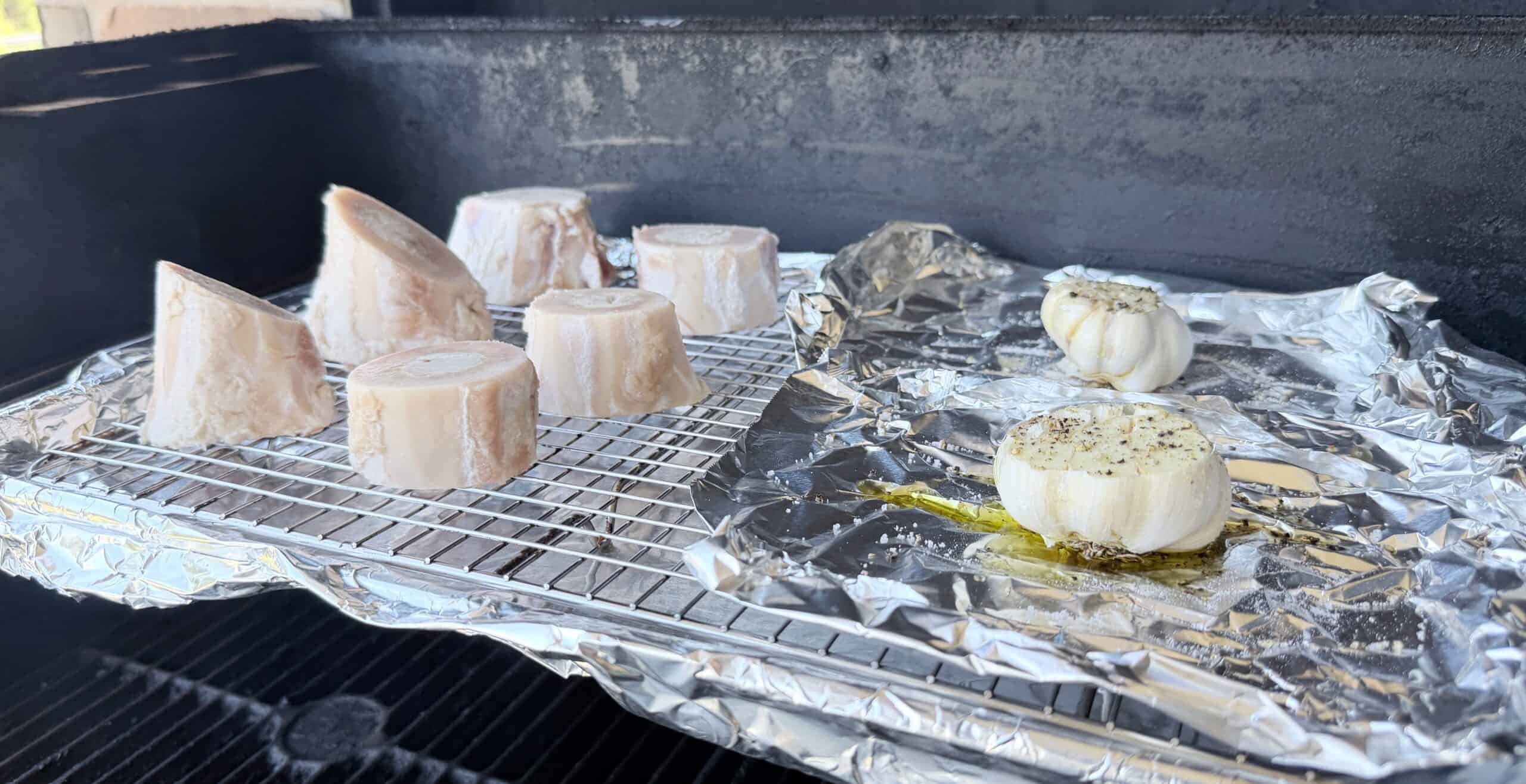Frozen marrow bones and two heads of garlic drizzled with oil and seasoning are placed on a rack lined with foil, inside a grill or smoker.