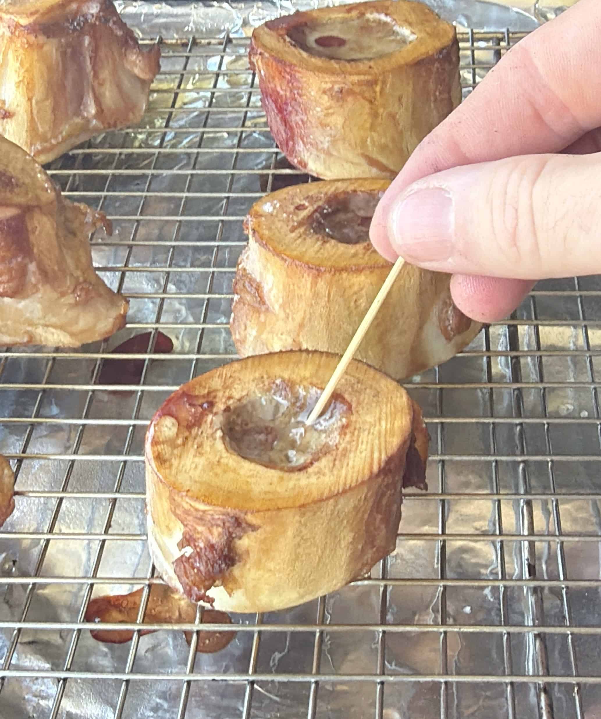 A hand uses a toothpick to scoop roasted bone marrow from a cross-sectioned bone sitting on a wire rack over a foil-lined tray. Other pieces of roasted bone are visible in the background.