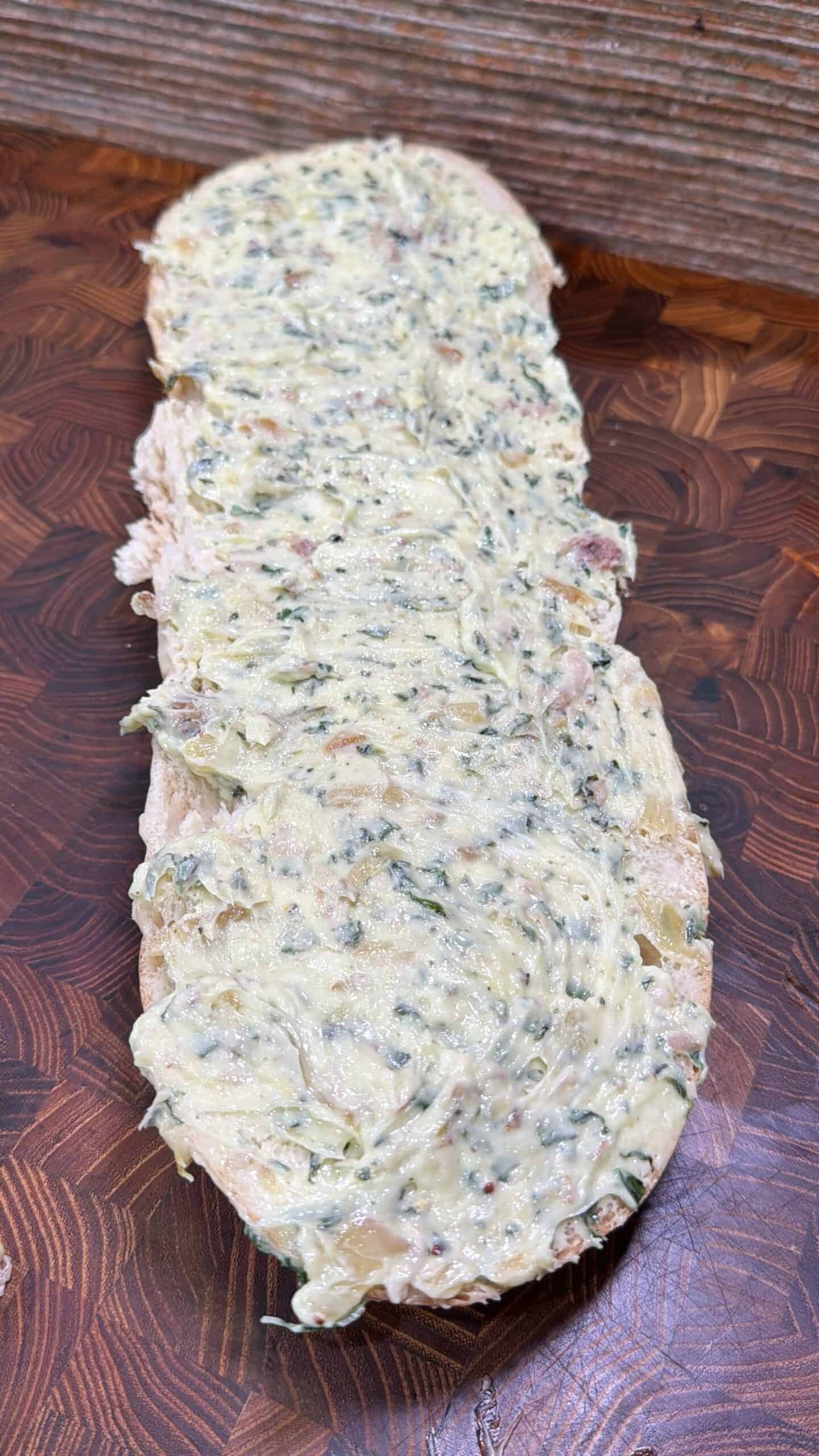 A slice of rustic bread topped with a thick layer of herb and garlic butter, placed on a patterned wooden cutting board.