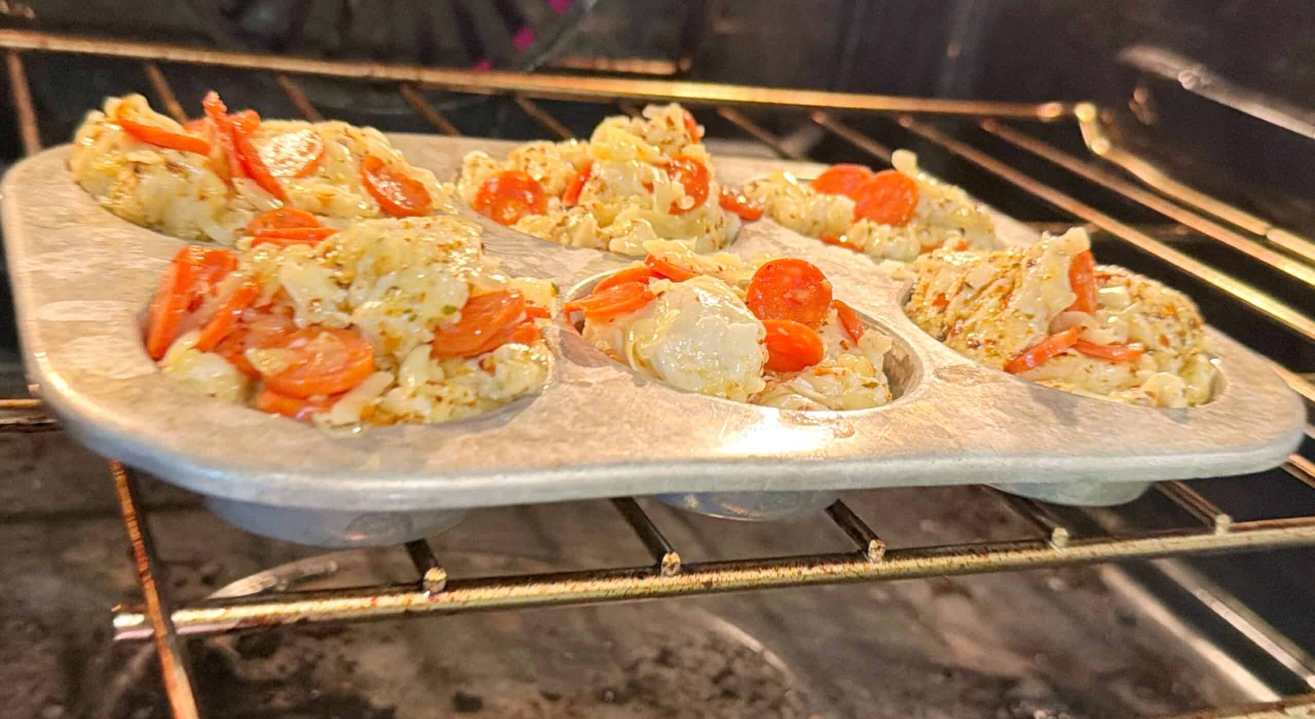 A metal muffin tin in an oven contains uncooked pizza muffin batter topped with mini pepperoni slices and cheese, set to bake.