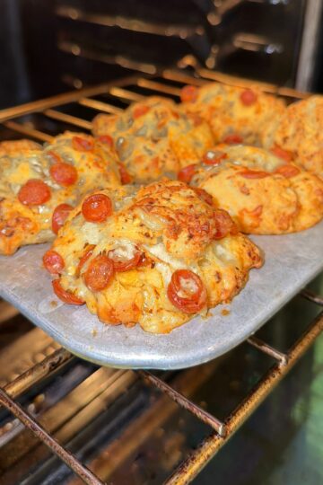 A baking tray with several freshly baked pizza-inspired biscuits topped with melted cheese and mini pepperoni, inside an oven.