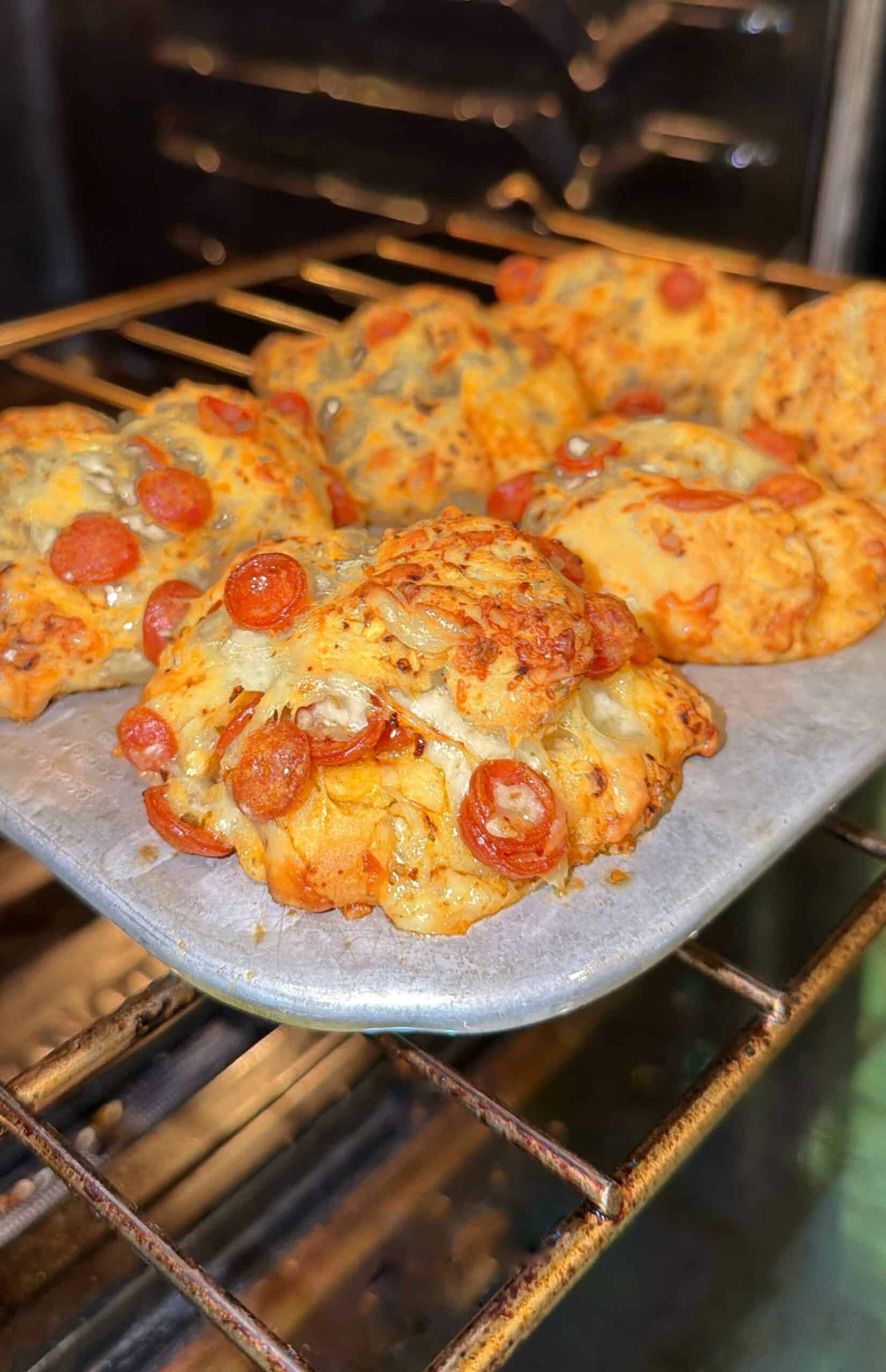 A baking tray with several freshly baked pizza-inspired biscuits topped with melted cheese and mini pepperoni, inside an oven.