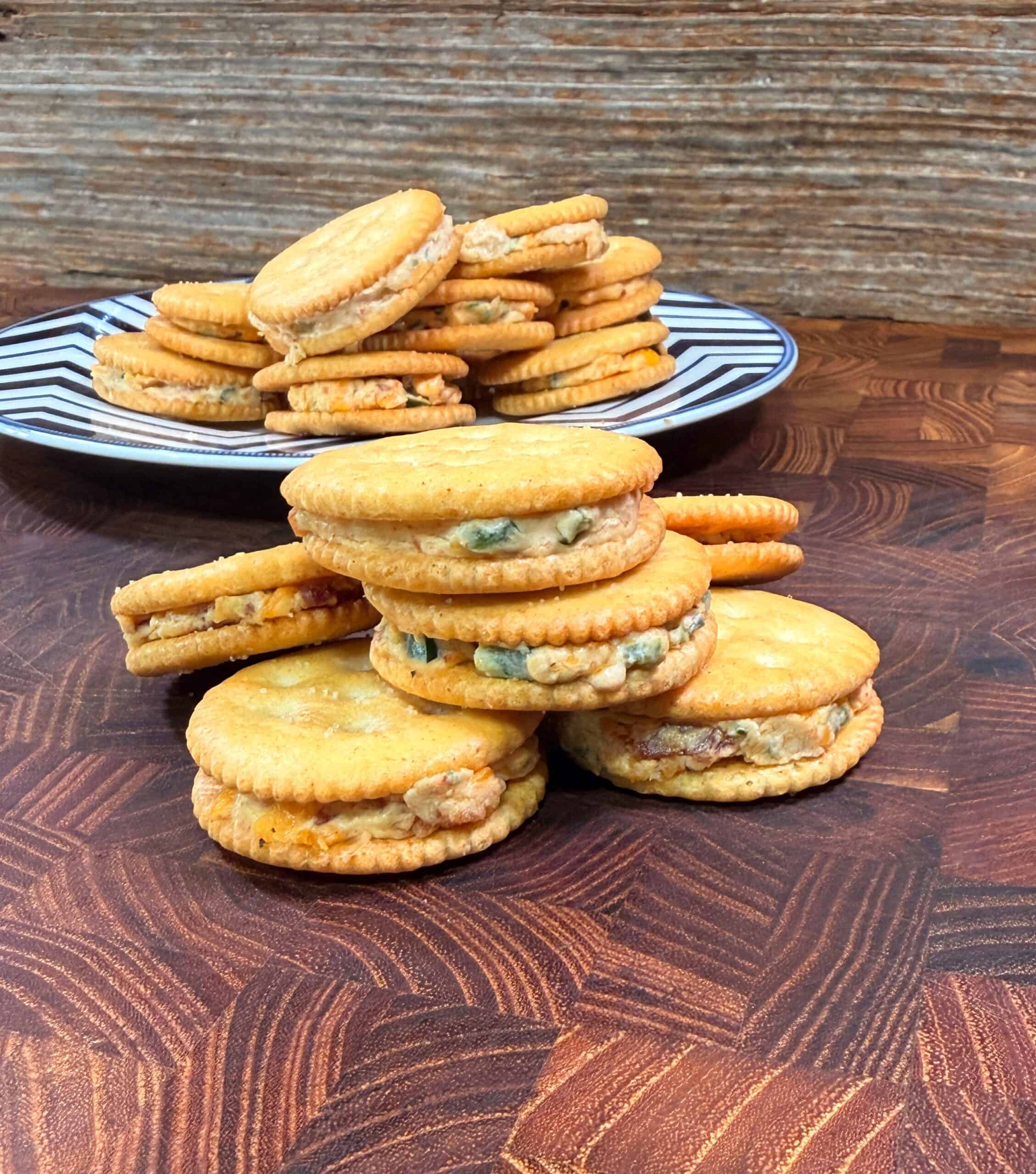 A close-up of sandwich crackers with various creamy fillings stacked on a wooden surface, with more crackers on a striped plate in the background against a wooden backdrop.