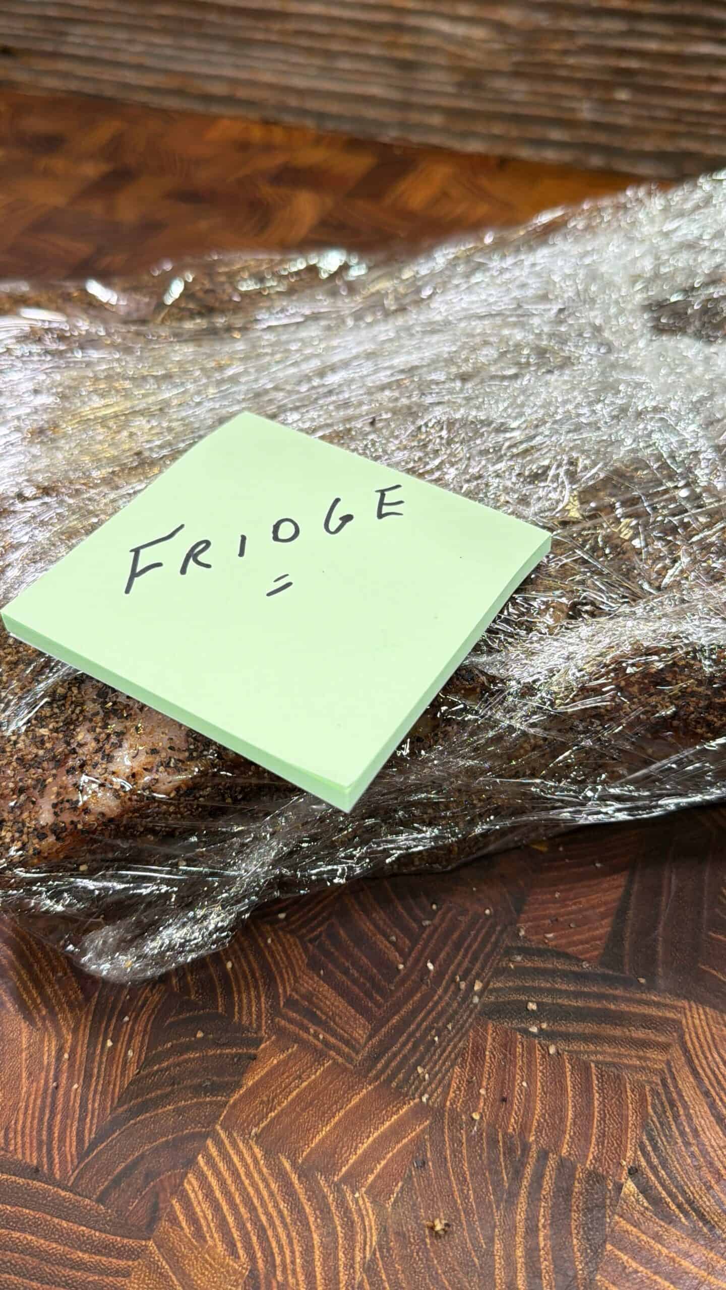 A piece of meat wrapped in plastic wrap sits on a wooden surface. A green sticky note labeled Fridge with a misspelling is placed on top.